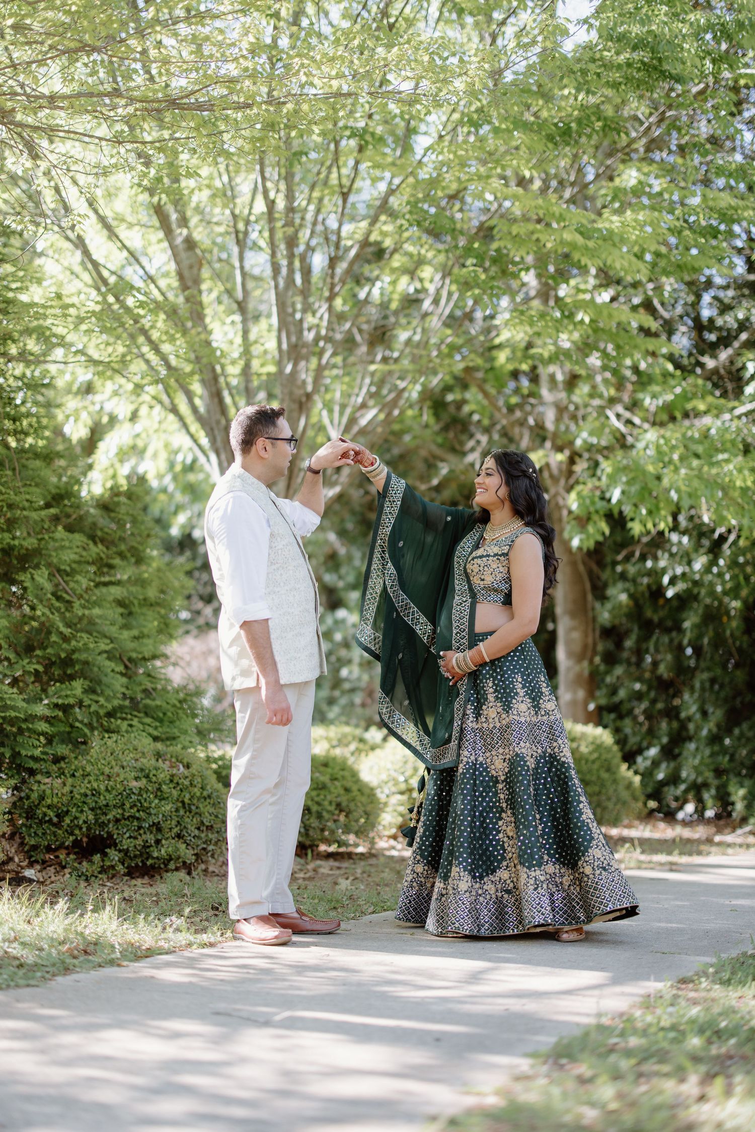 A couple in traditional and fusion attire share a romantic moment on a pathway surrounded by lush greenery.