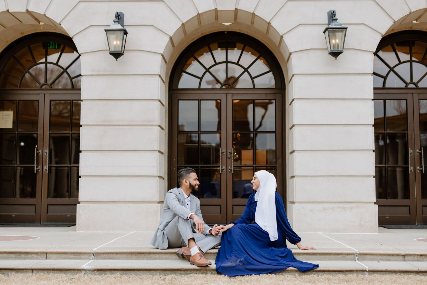 Two people sit on stone steps in front of an elegant building with arched doorways and lanterns.