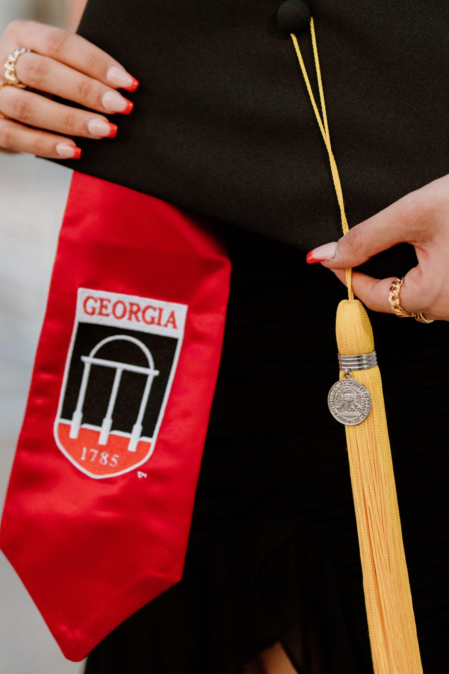 Graduate holding yellow tassel and UGA graduation stole with red Georgia arch logo against black background.