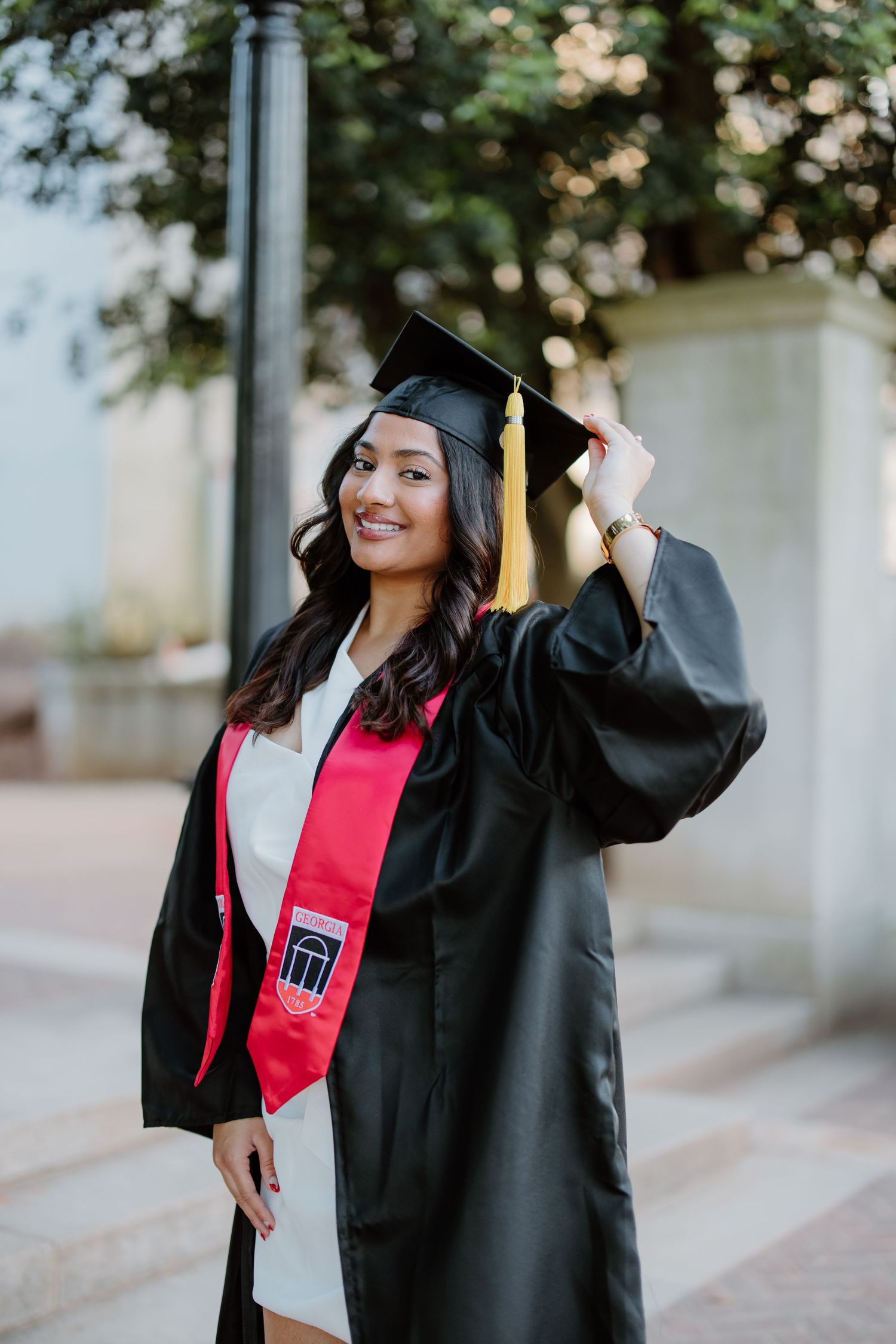 A graduate wearing a traditional black cap and gown with red stole stands outside celebrating their commencement.