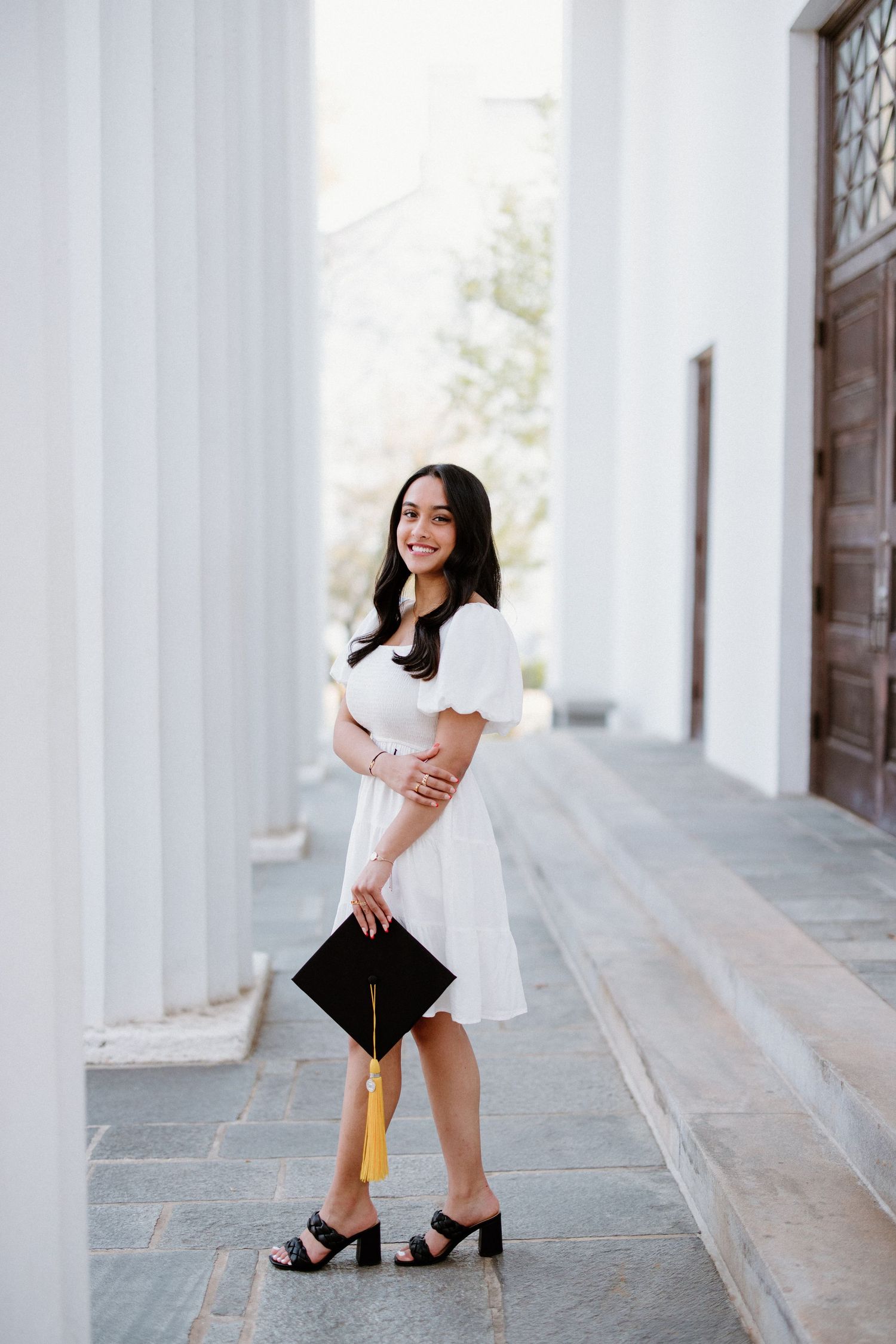 Woman in white dress and black heels holds graduation cap while posing on stone steps near columns.
