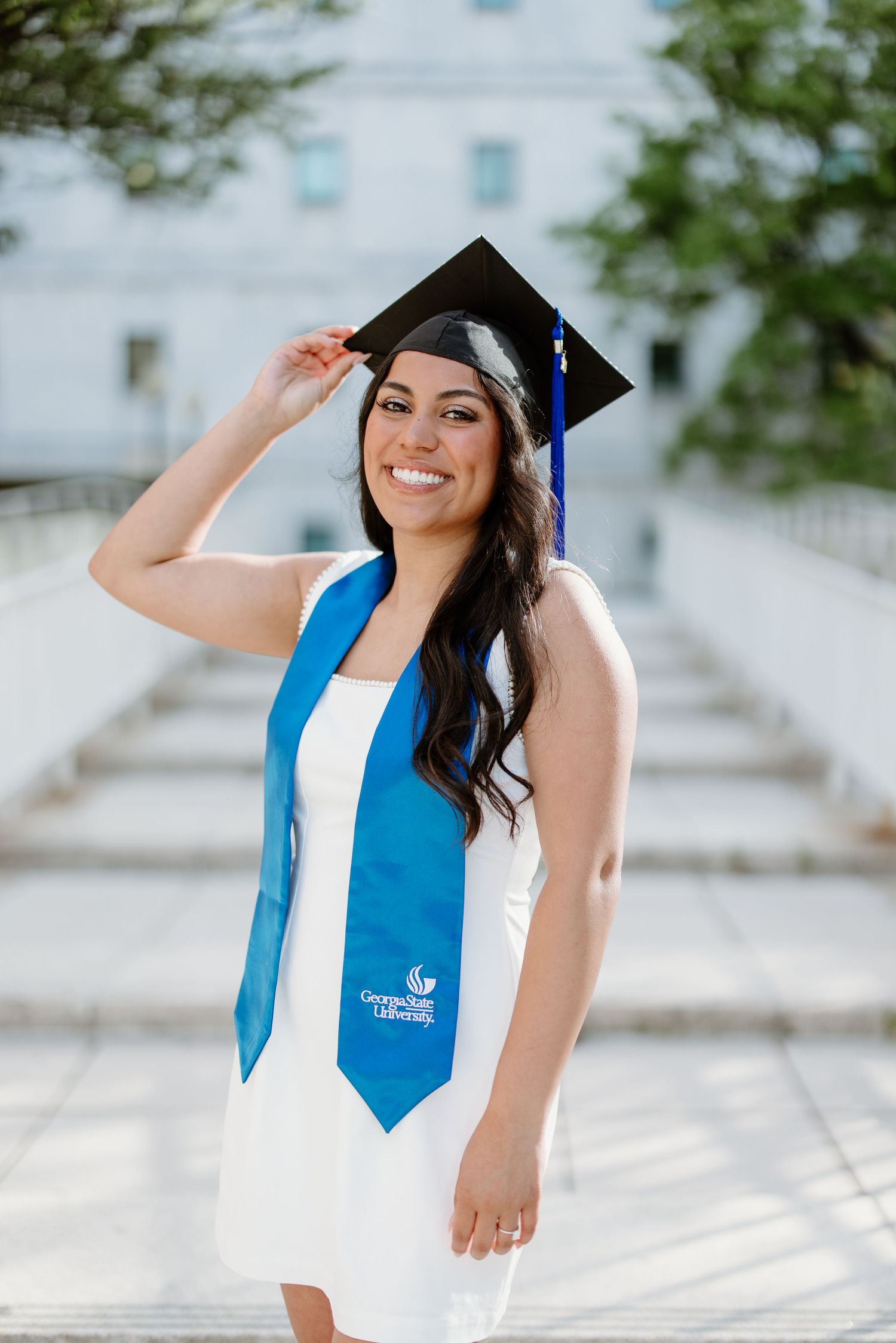 Graduate in white dress and blue graduation stole poses outdoors for graduation photos on sunny day.