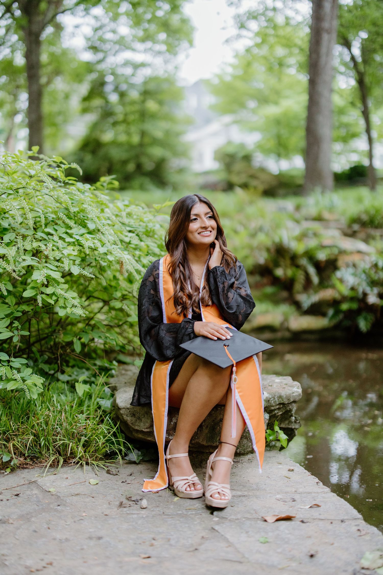 A graduate in a black gown and yellow dress sits on a rock by a peaceful stream in a lush green garden setting.