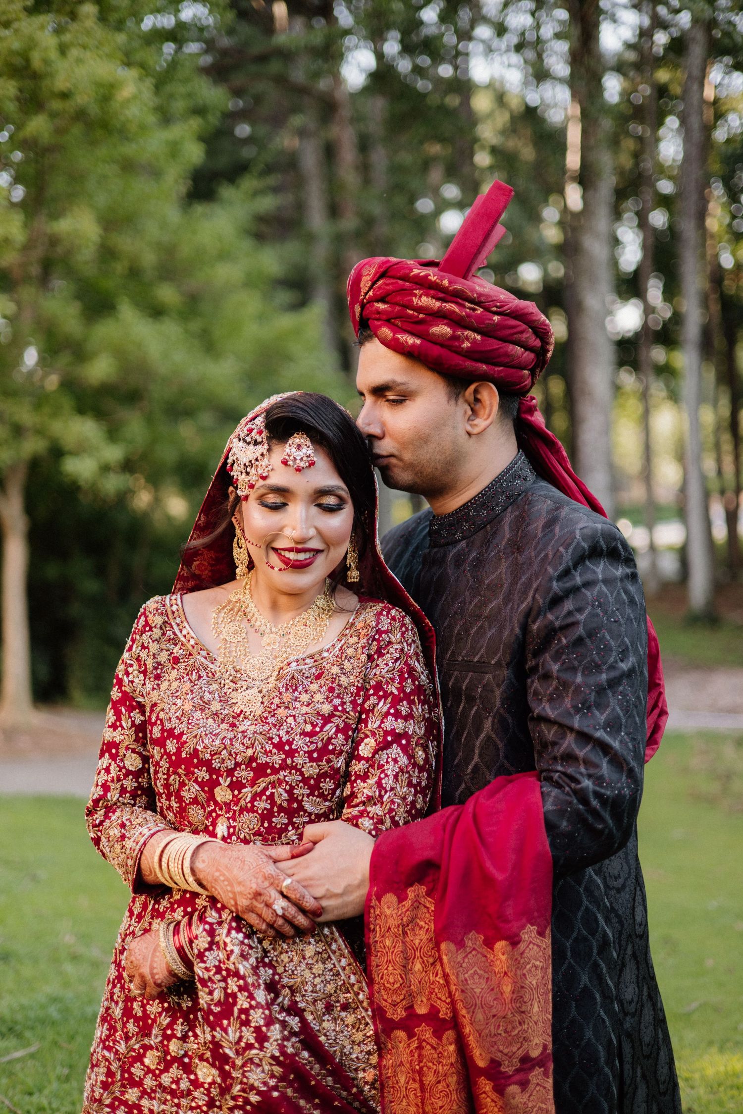 Traditional South Asian couple embraces in a park wearing elegant red and black wedding attire.