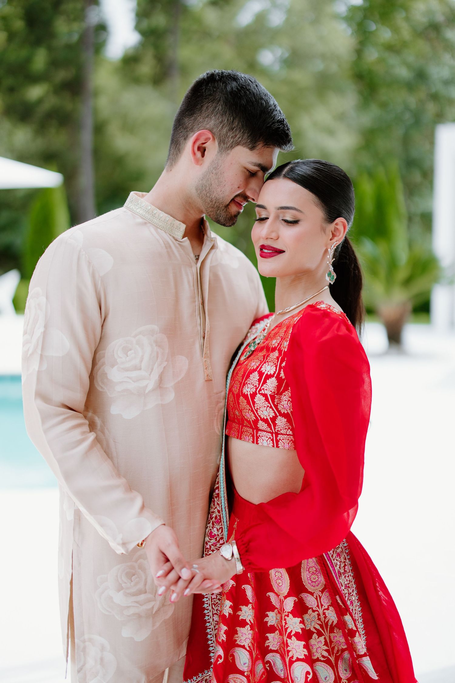 A couple in traditional South Asian wedding attire embrace, with the bride in red lehenga and groom in beige kurta.