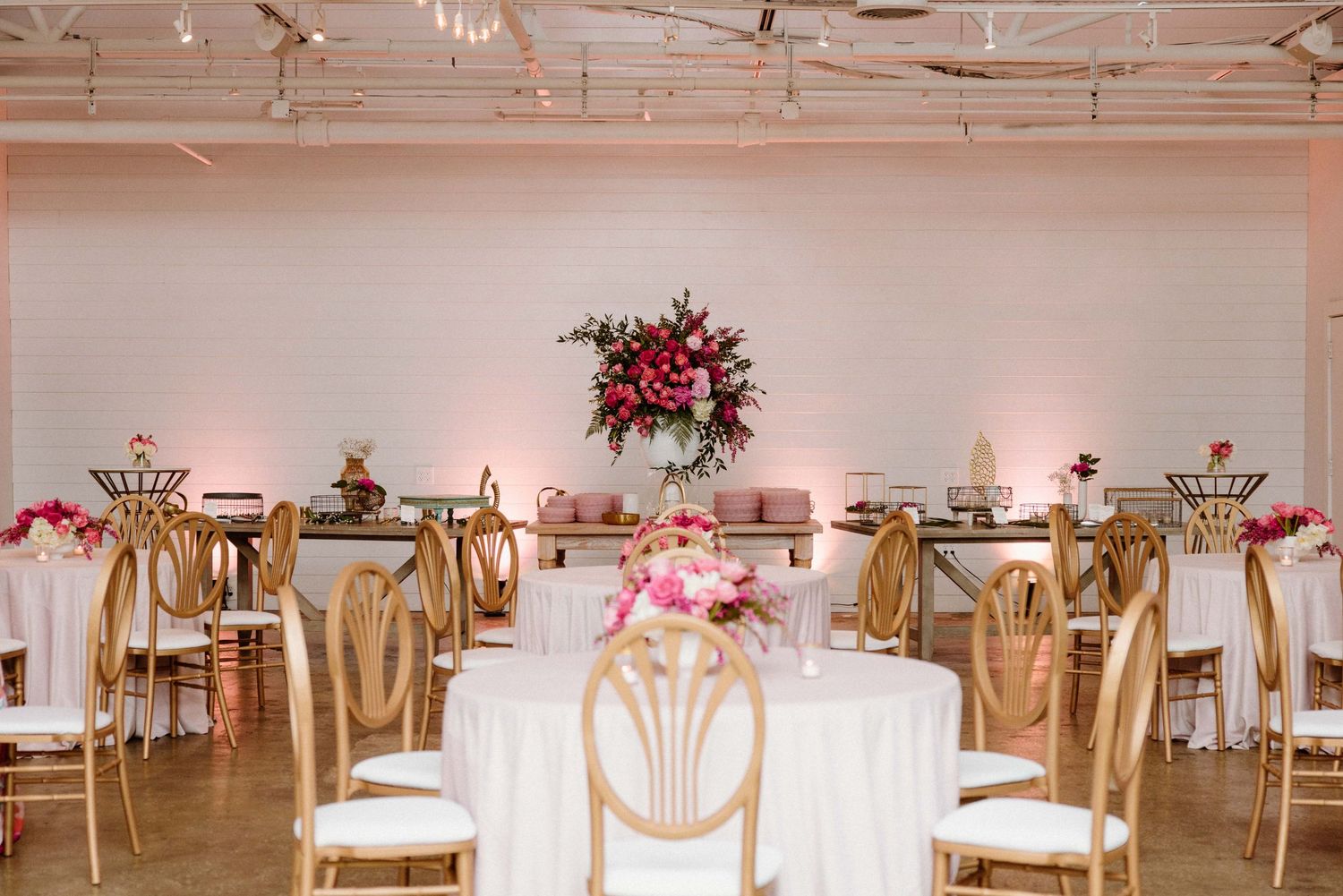 Elegant wedding reception setup with white tables, gold chair sashes, and pink floral arrangements in an indoor venue.