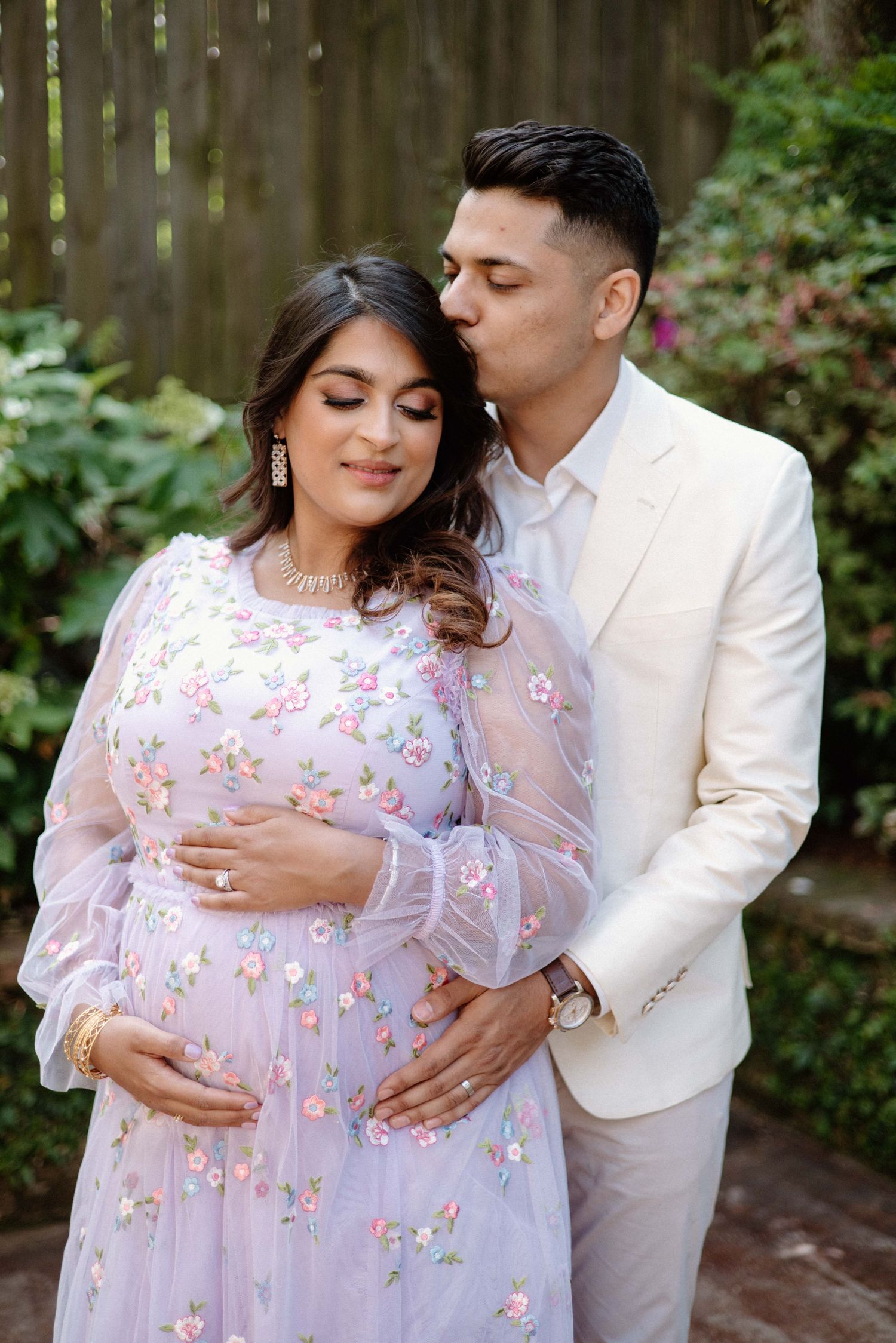 A couple shares an intimate moment outdoors, with the partner in white embracing the other wearing a flowing floral dress.