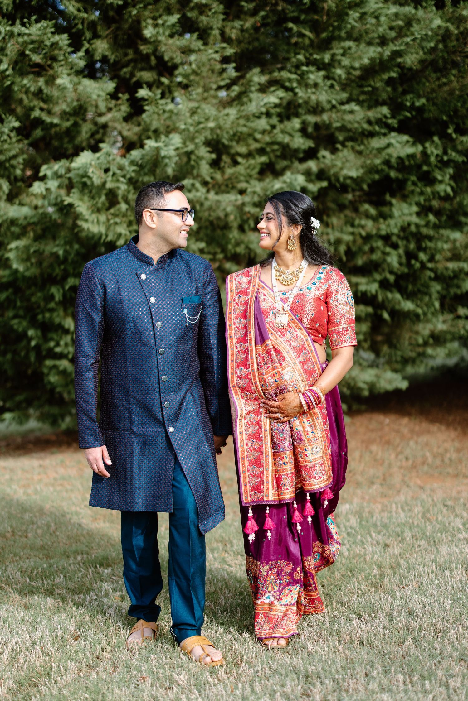 A couple wearing traditional fusion wedding attire, with navy blue sherwani and pink floral sari, stand together outdoors.