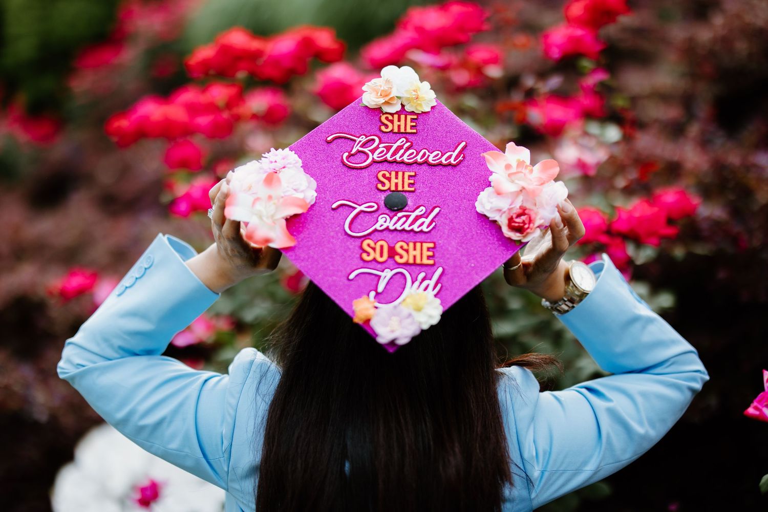 View from behind of someone in light blue jacket holding up a pink graduation cap among blooming pink flowers.