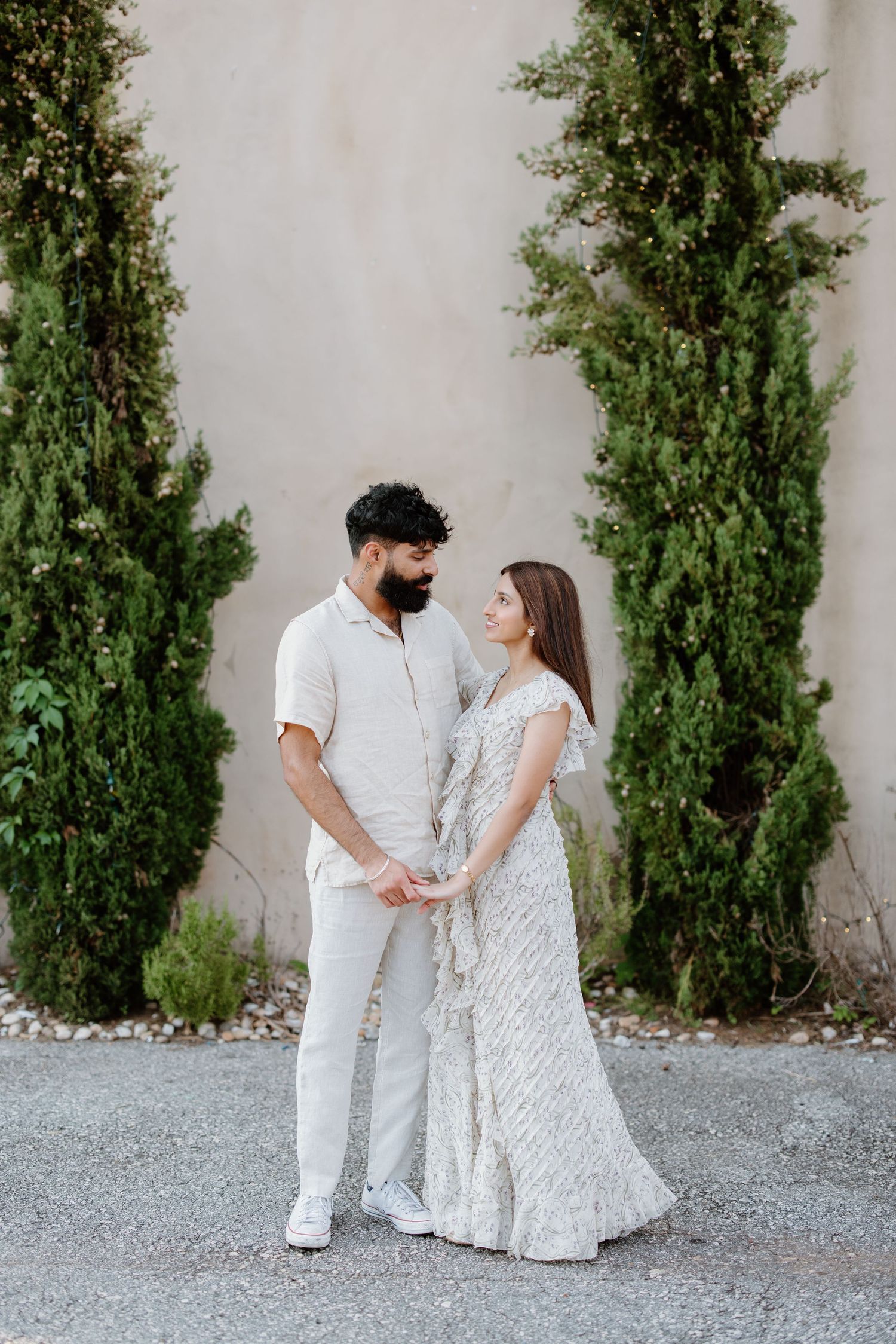 Couple in elegant all-white outfits pose together against stone wall with tall cypress trees in romantic outdoor setting.