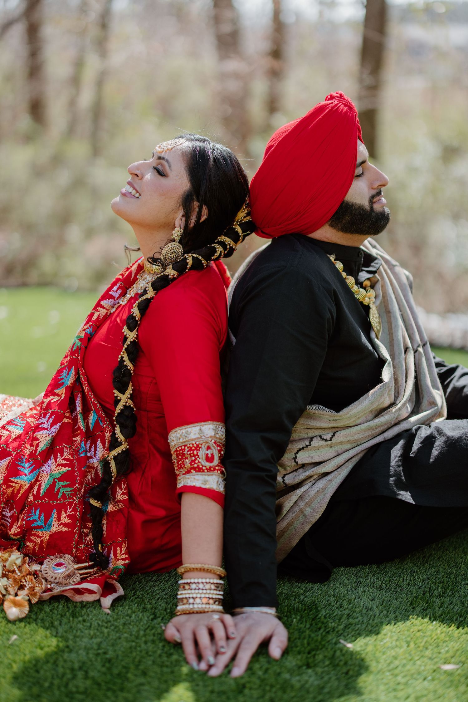 A couple in traditional South Asian attire sitting back to back outdoors, wearing red and black clothing with ornate details.