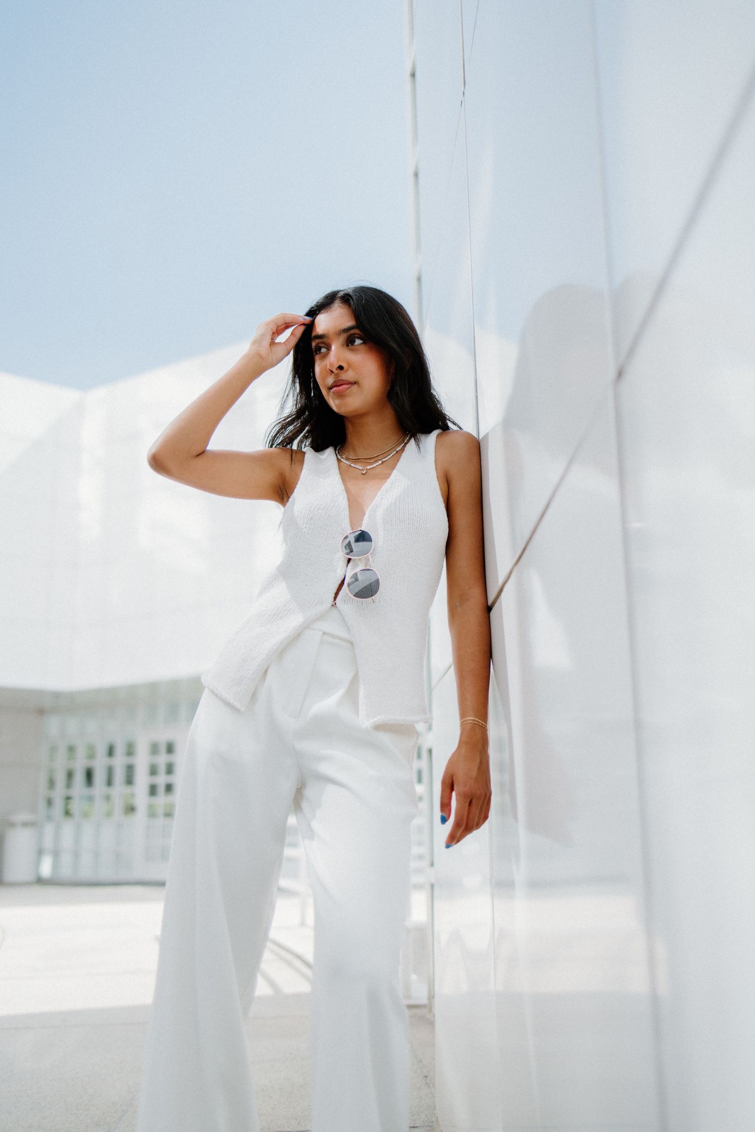 A person in an all-white outfit stands against white curtains in a bright, minimalist setting with sunglasses hanging on the neckline.
