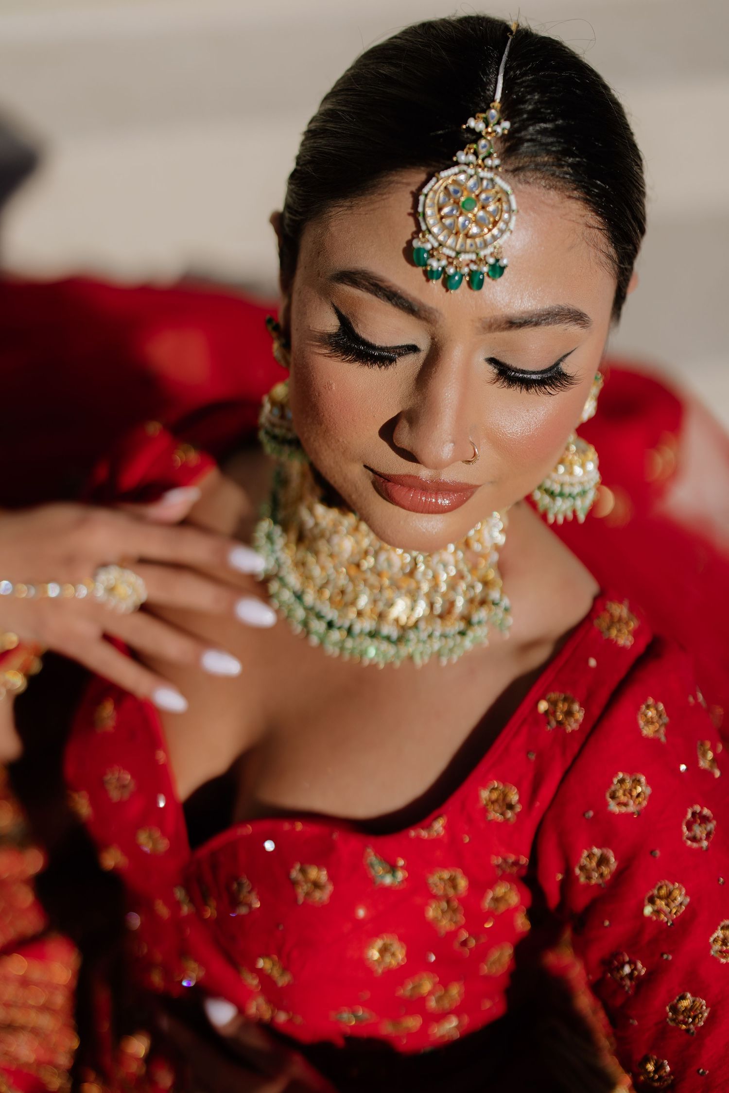 A bride adorned in traditional Indian wedding jewelry and red attire displays intricate makeup and ornate accessories.