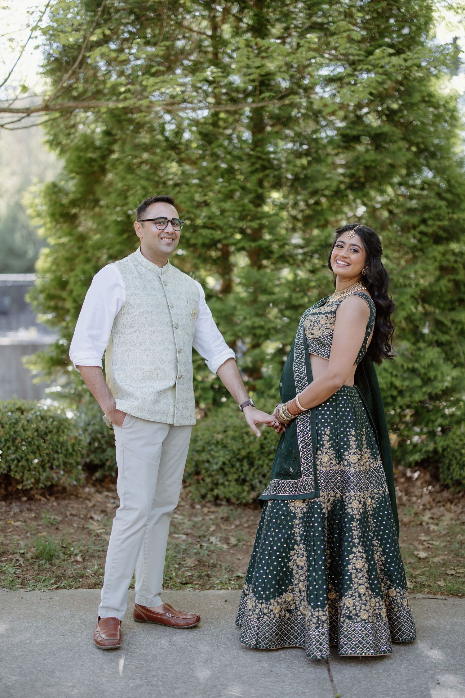 A couple in elegant formal wear standing together on a garden path with trees in the background.