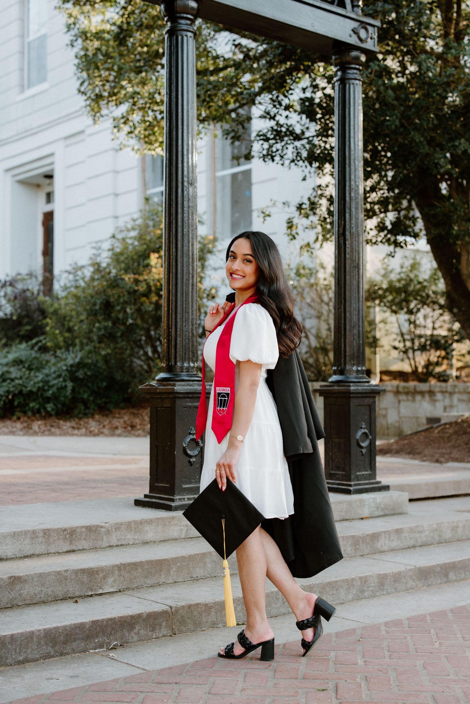 Graduate in black graduation gown and hood poses with stoles on university campus steps, celebrating achievement.