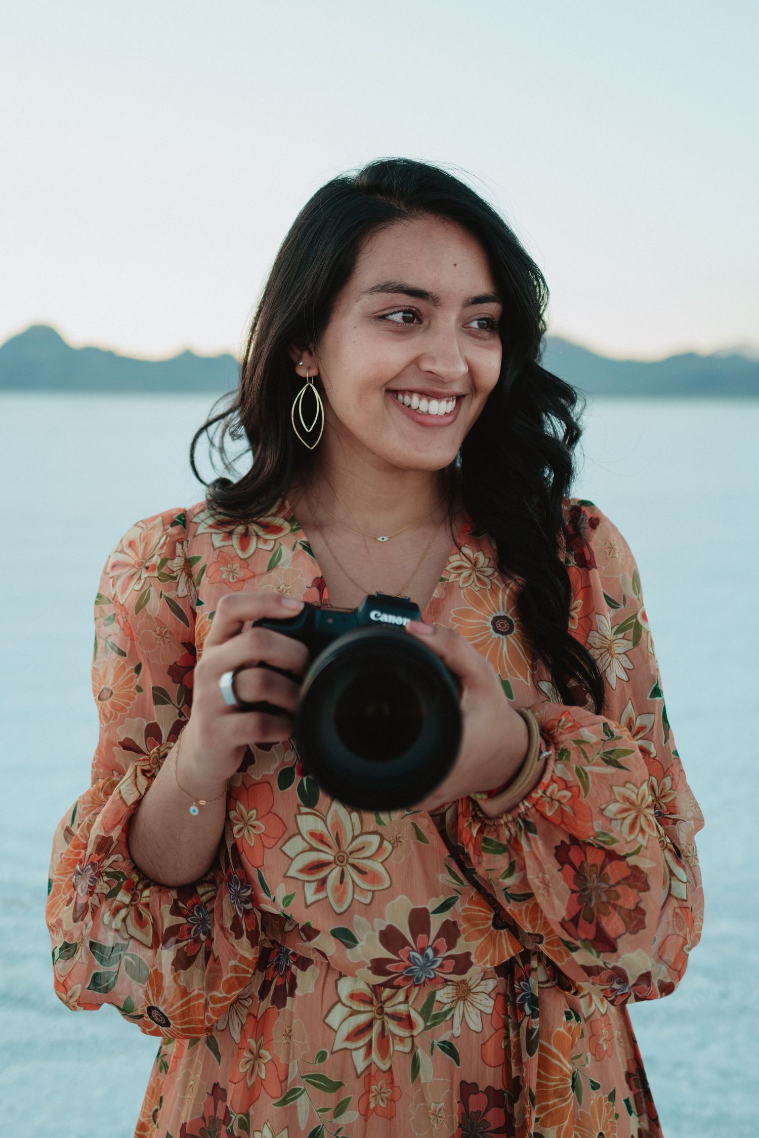 Photographer in floral orange dress smiles while holding DSLR camera against serene waterfront background.