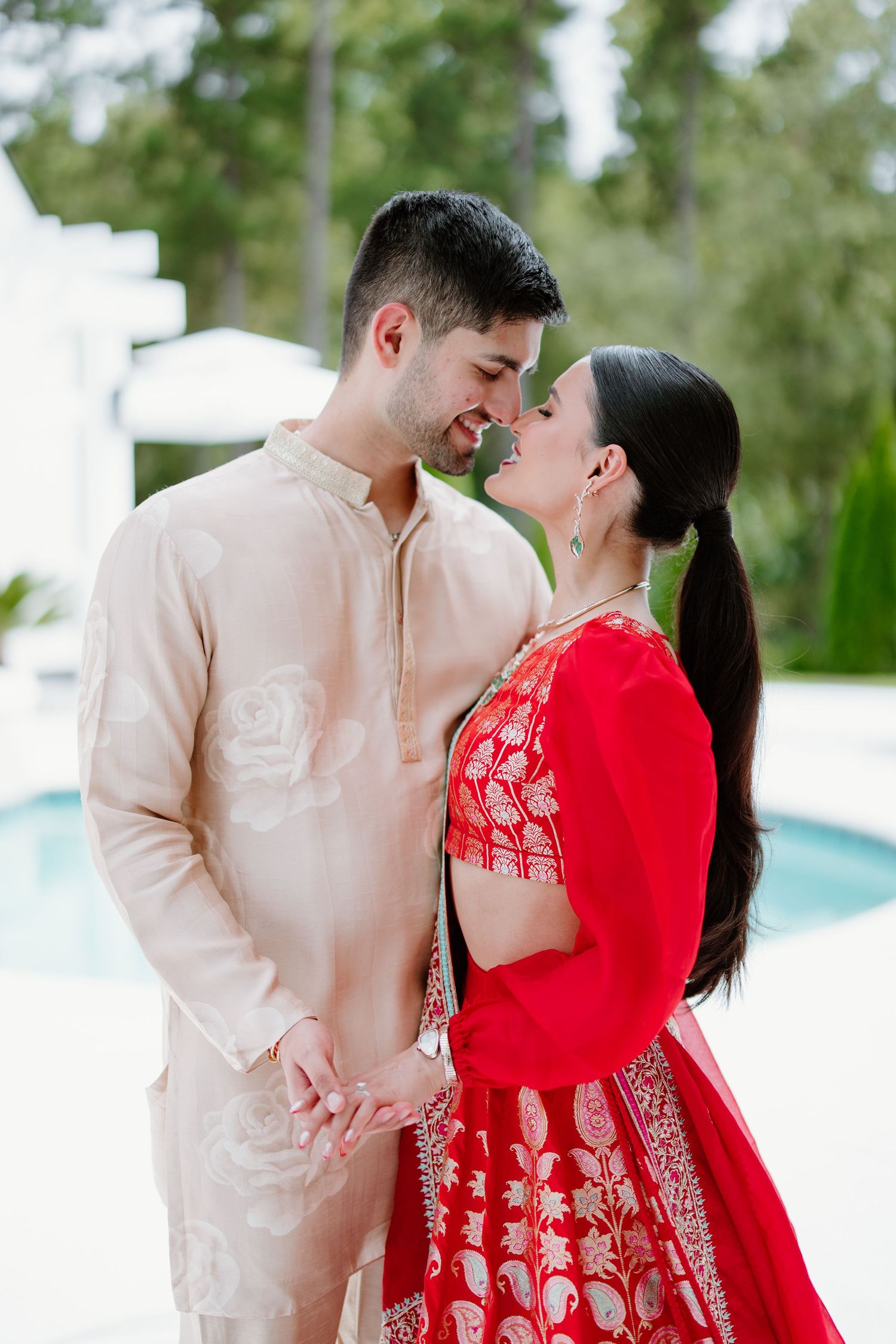 A couple in coordinating red and beige traditional Asian formal wear share romantic moments near a pool.