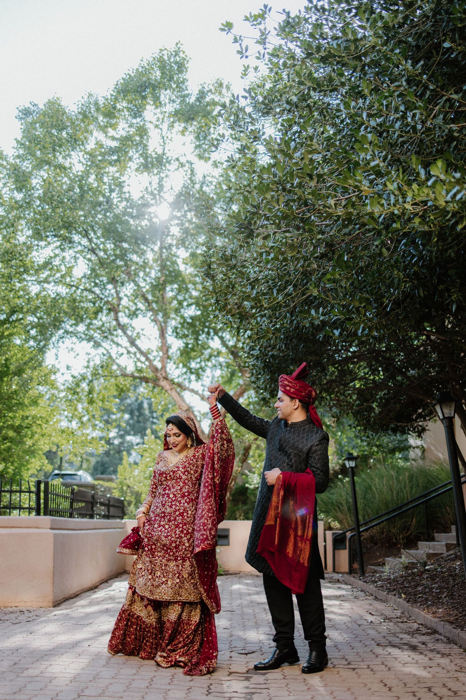 Two people in traditional South Asian wedding attire dance together on a pathway surrounded by green trees.