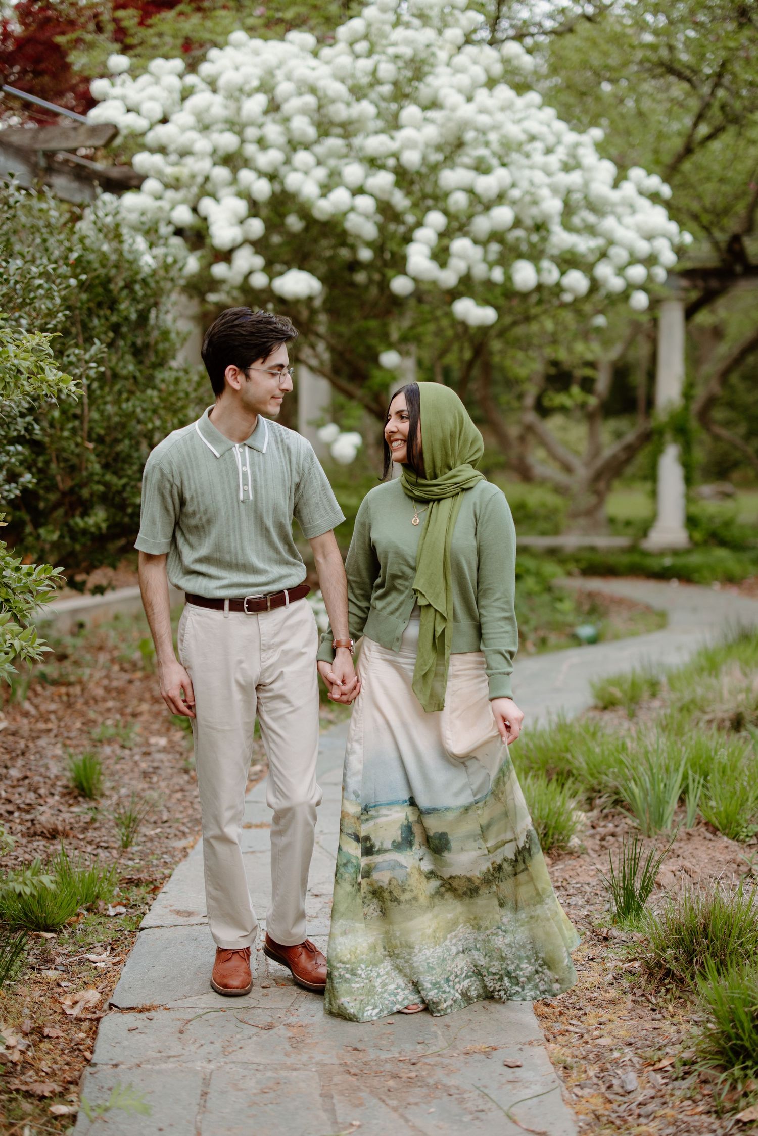 A couple holding hands walks along a garden path under blooming white flowering trees in springtime.