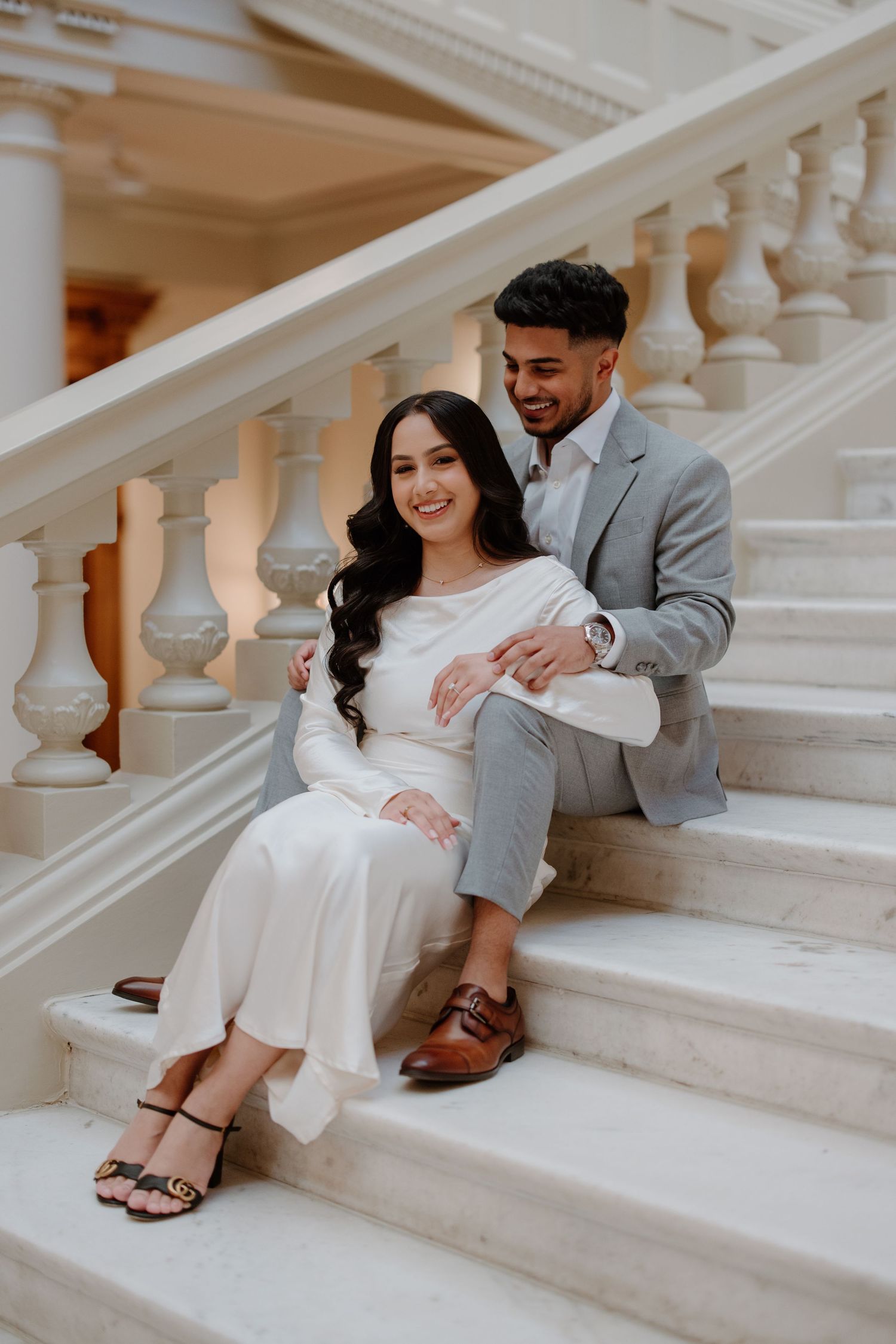 A couple in formal attire sitting together on elegant white marble stairs in a romantic engagement photo session.