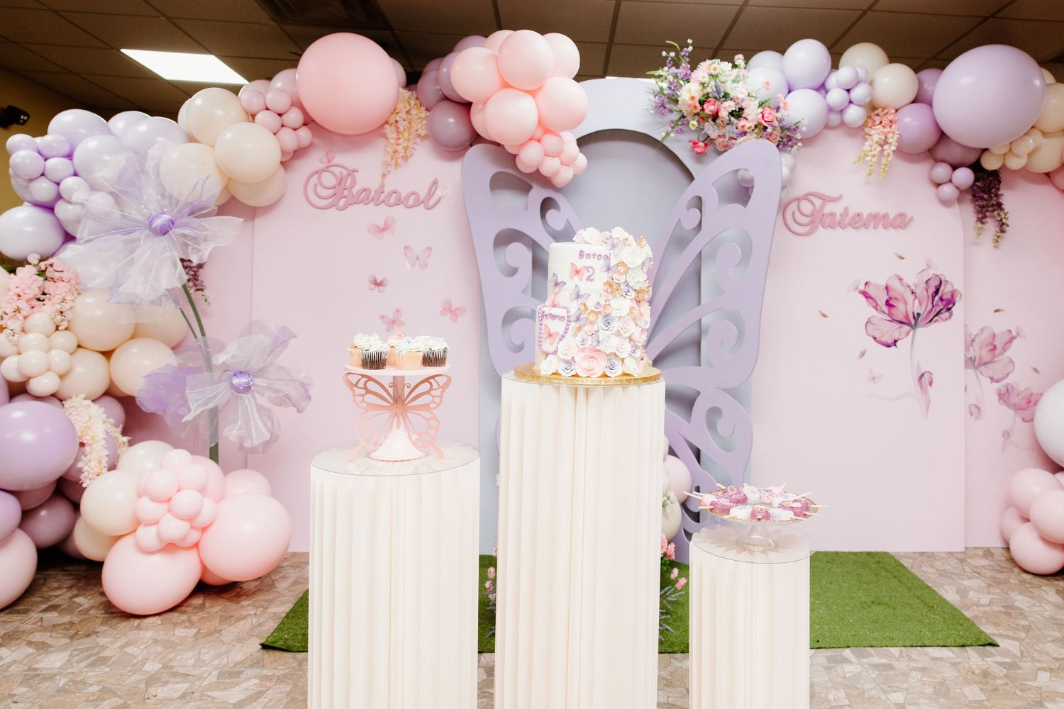 Elegant party setup with pink and purple balloon arch, white pedestals, and butterfly decor against a pastel backdrop.