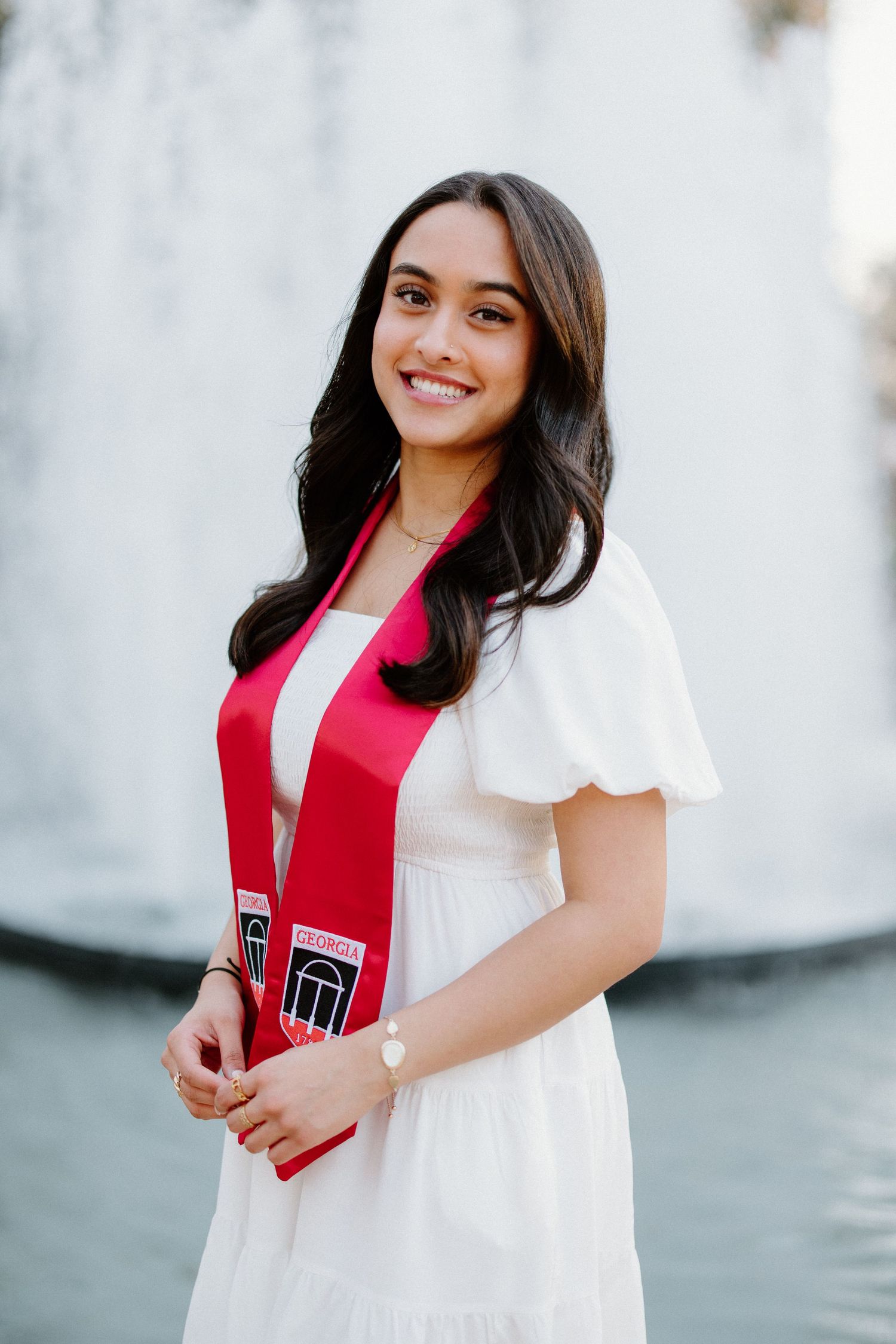 A graduate in a white dress and red graduation sash smiles brightly in an outdoor portrait against a light wall.