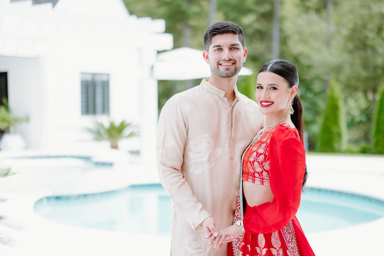 Wedding couple in traditional Indian attire poses together by pool with bride in vibrant red lehenga.