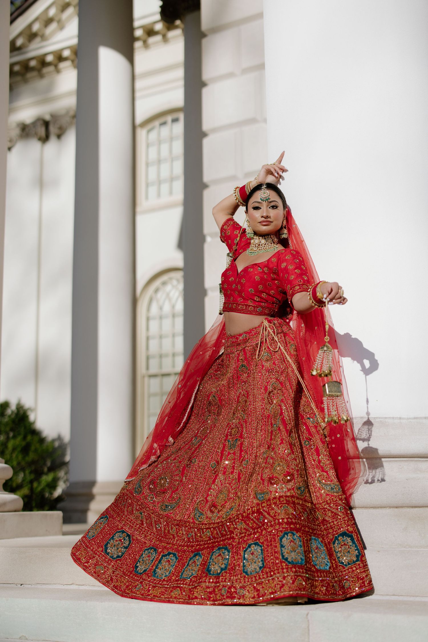 A dancer in a traditional red lehenga with intricate embroidery poses elegantly against a white columned building.