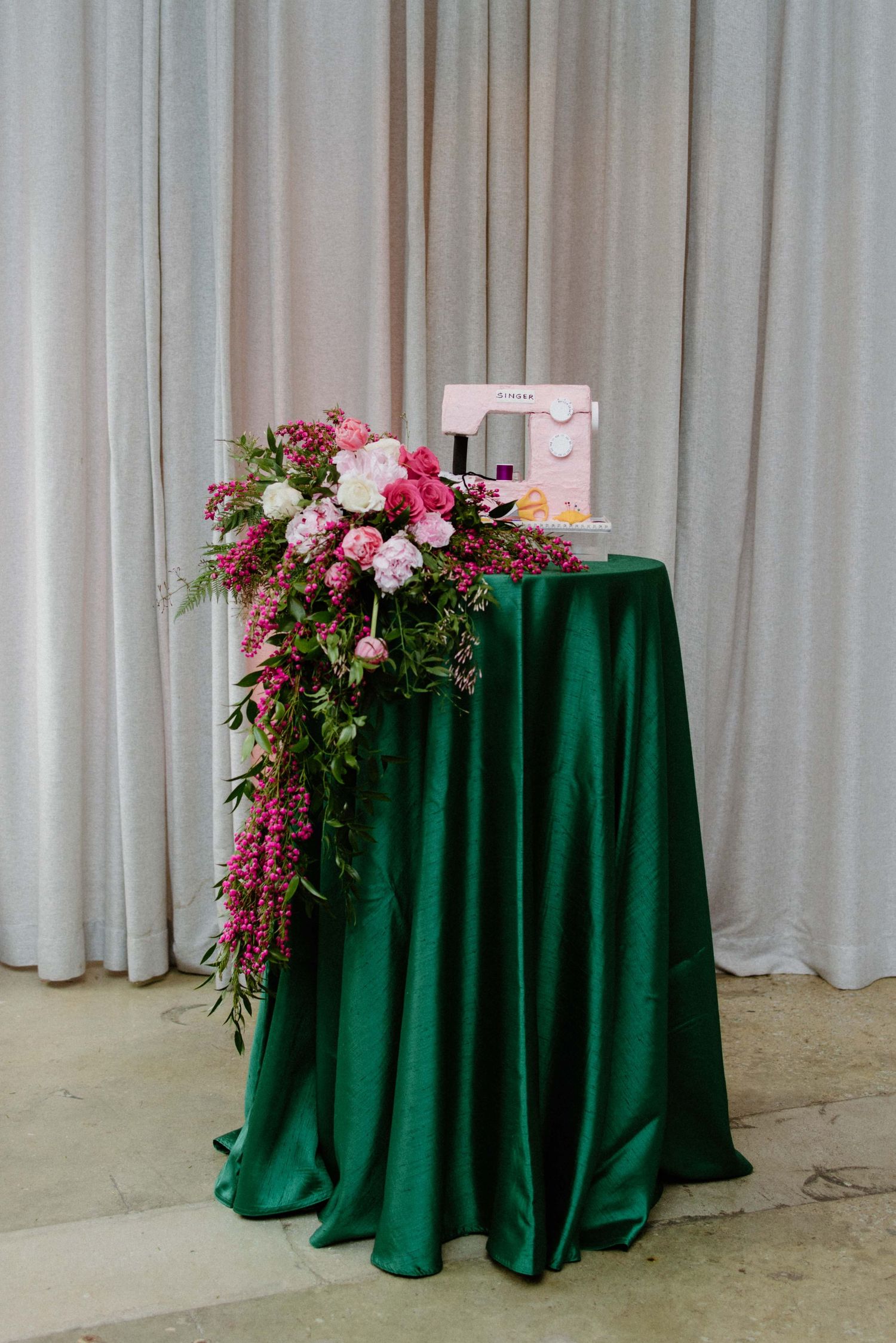 Pink floral arrangements adorn emerald green velvet tablecloths against a white curtain backdrop.