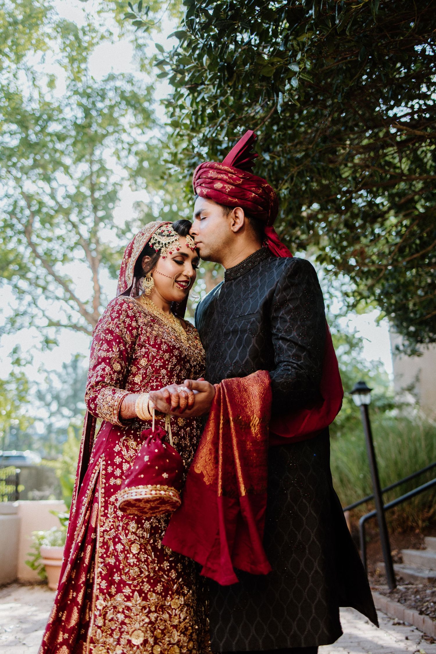 A couple in traditional South Asian wedding attire share an intimate moment outdoors among greenery and trees.