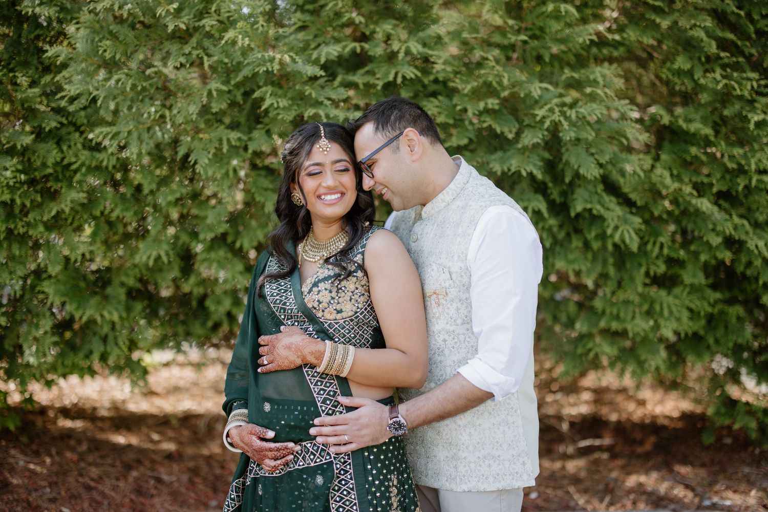 A romantic pregnancy photoshoot features a couple embracing in front of lush green foliage.