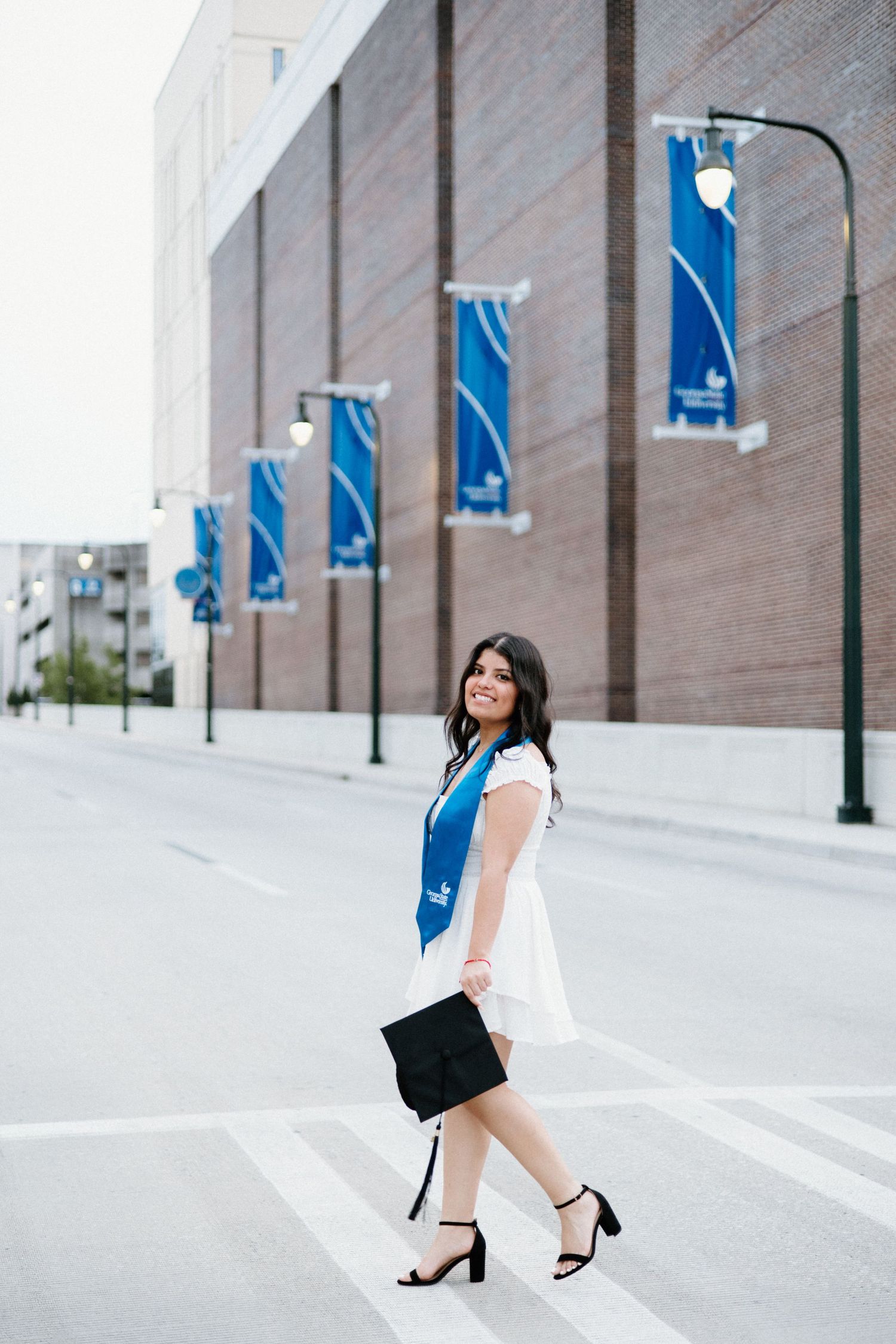Graduate in a white dress and blue sash walks down a city street with blue banners displayed along brick wall.