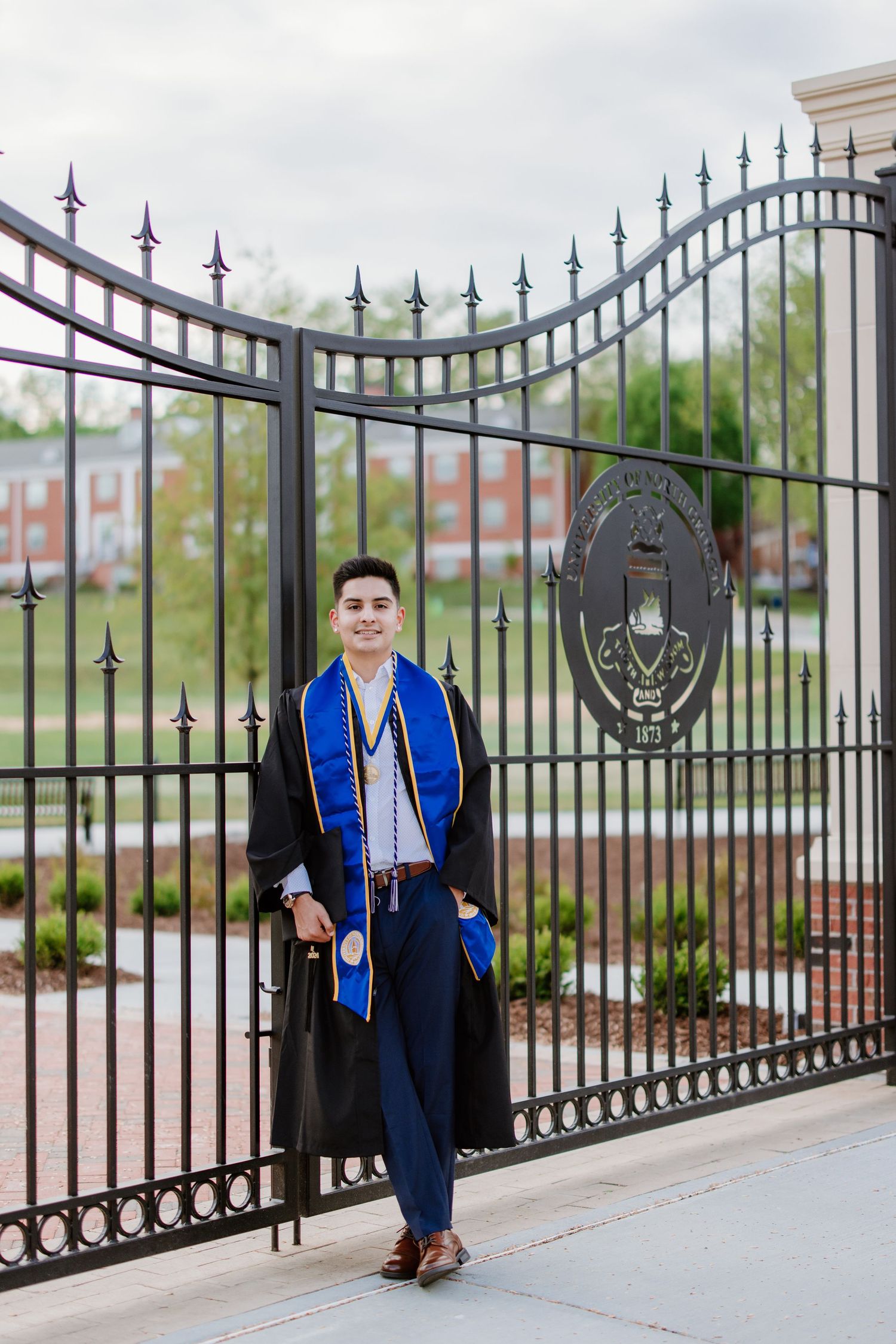 Graduate in cap and gown poses by ornate iron gates at a university campus with architecture visible in background.