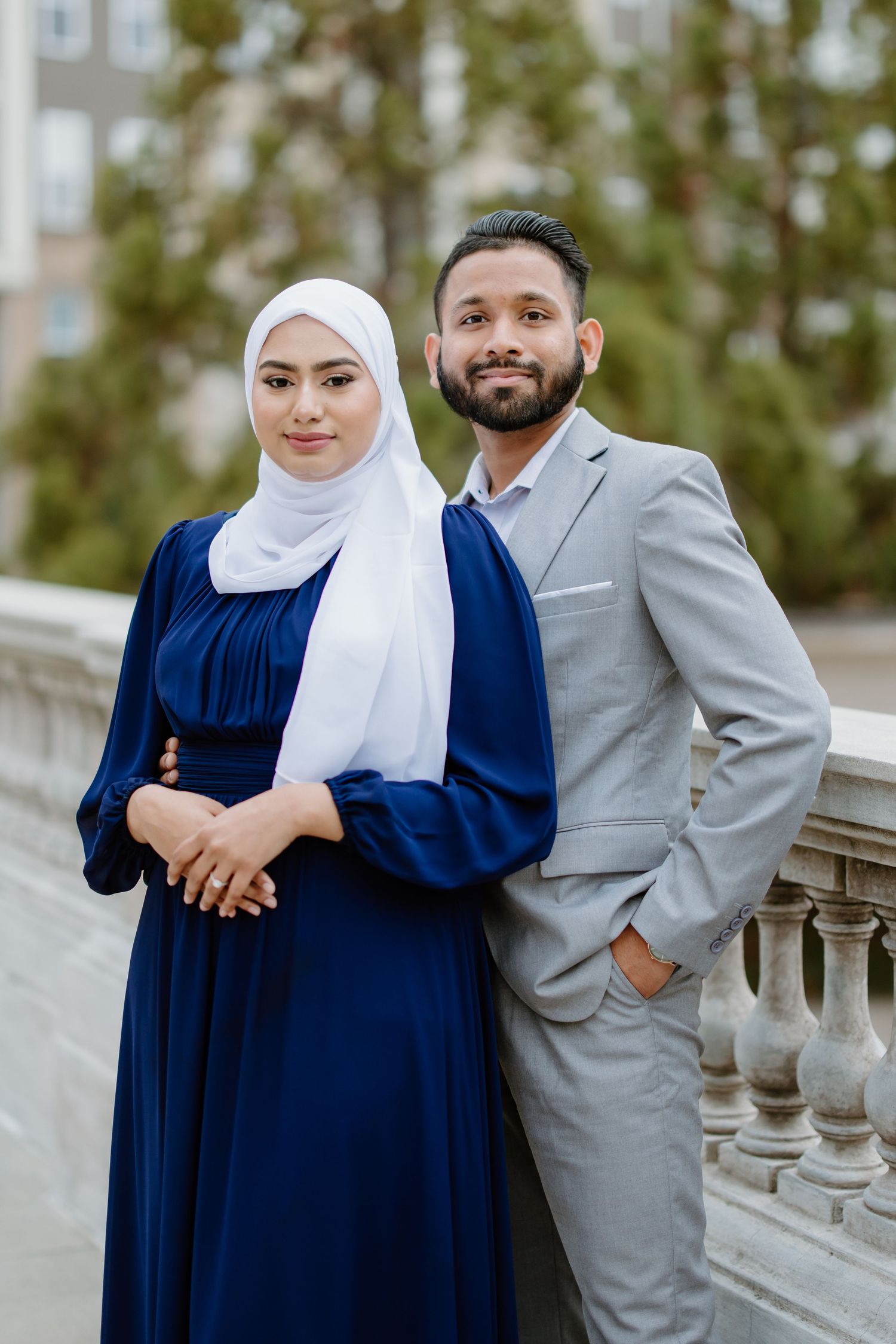 Couple standing together outdoors, one wearing a navy dress with white hijab and one in a light grey suit.