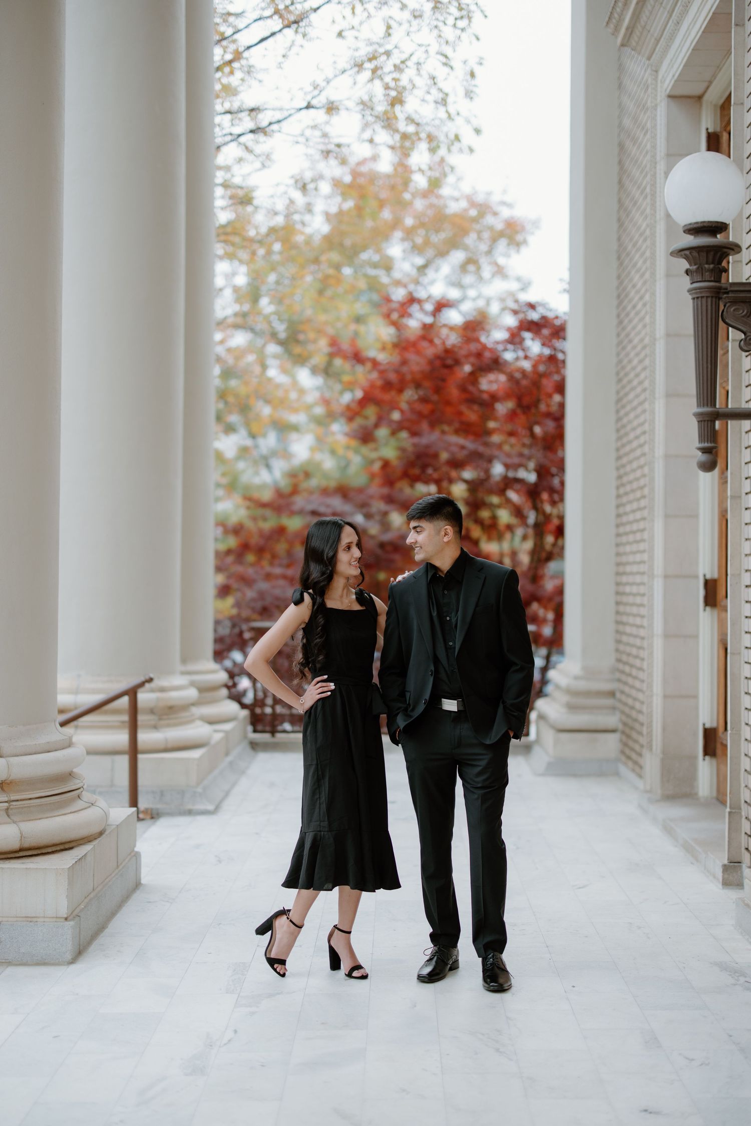 A couple in elegant black attire poses romantically between white columns with autumn foliage in the background.