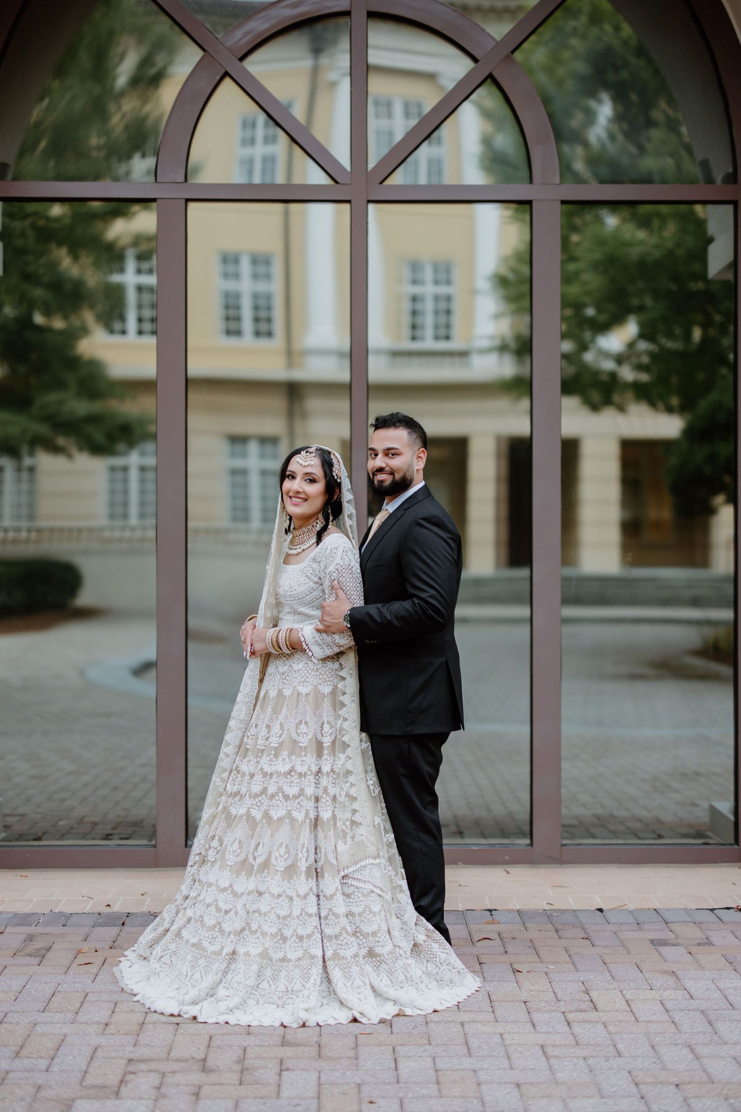 Newlyweds in formal attire share a romantic moment on a brick patio with elegant arched windows in the background.