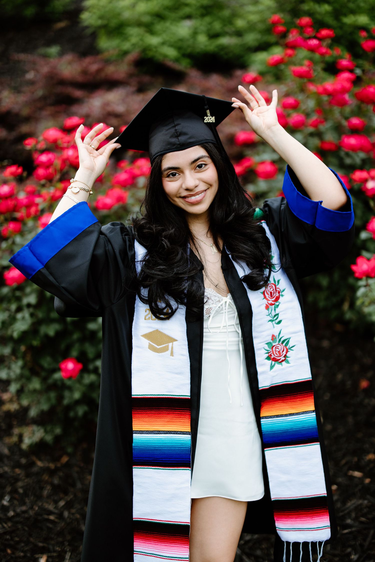 A graduate in cap and gown wearing a colorful serape-style graduation stole poses with raised arms against blooming red flowers.