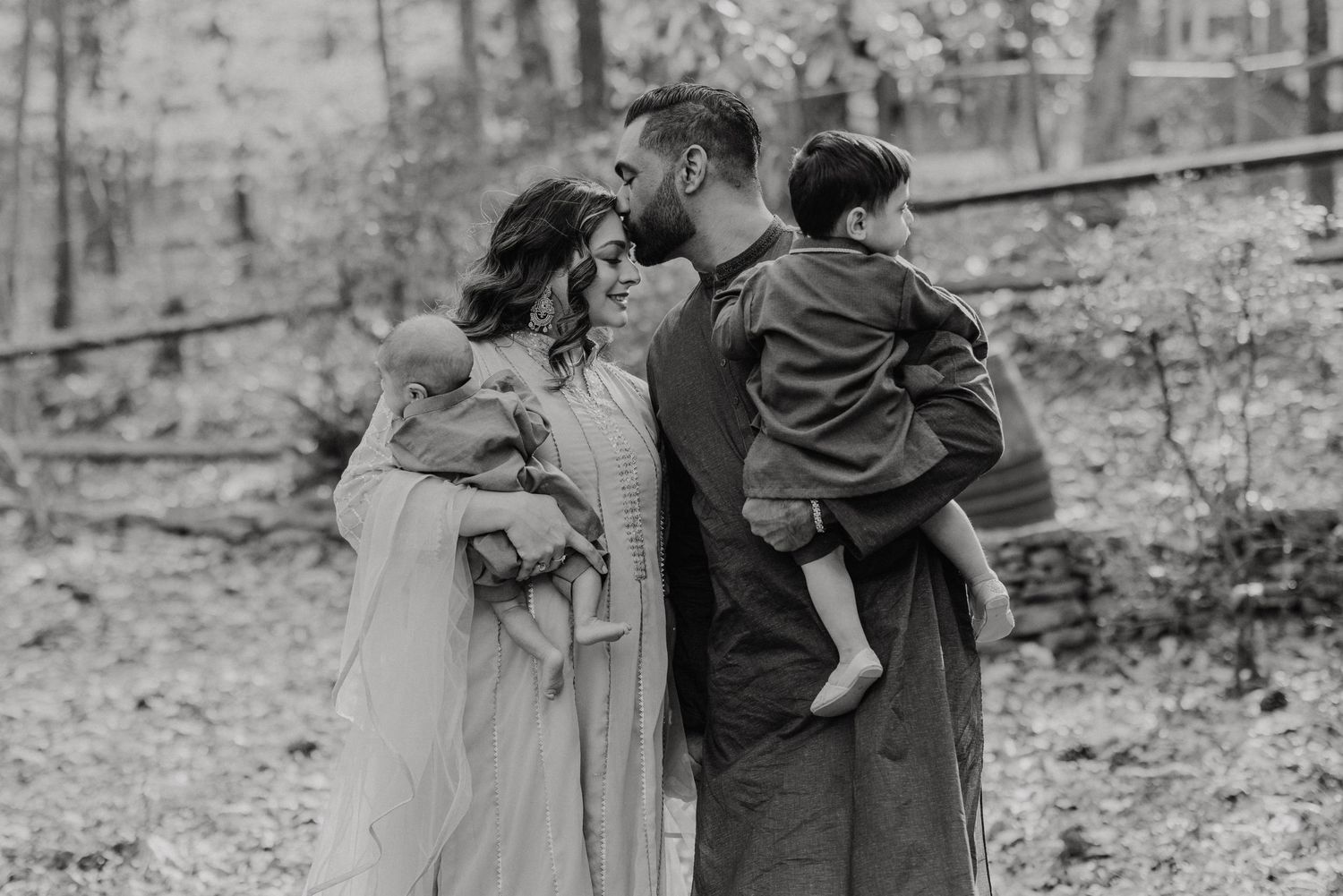 A family shares a tender moment together in black and white while standing in a wooded outdoor setting.