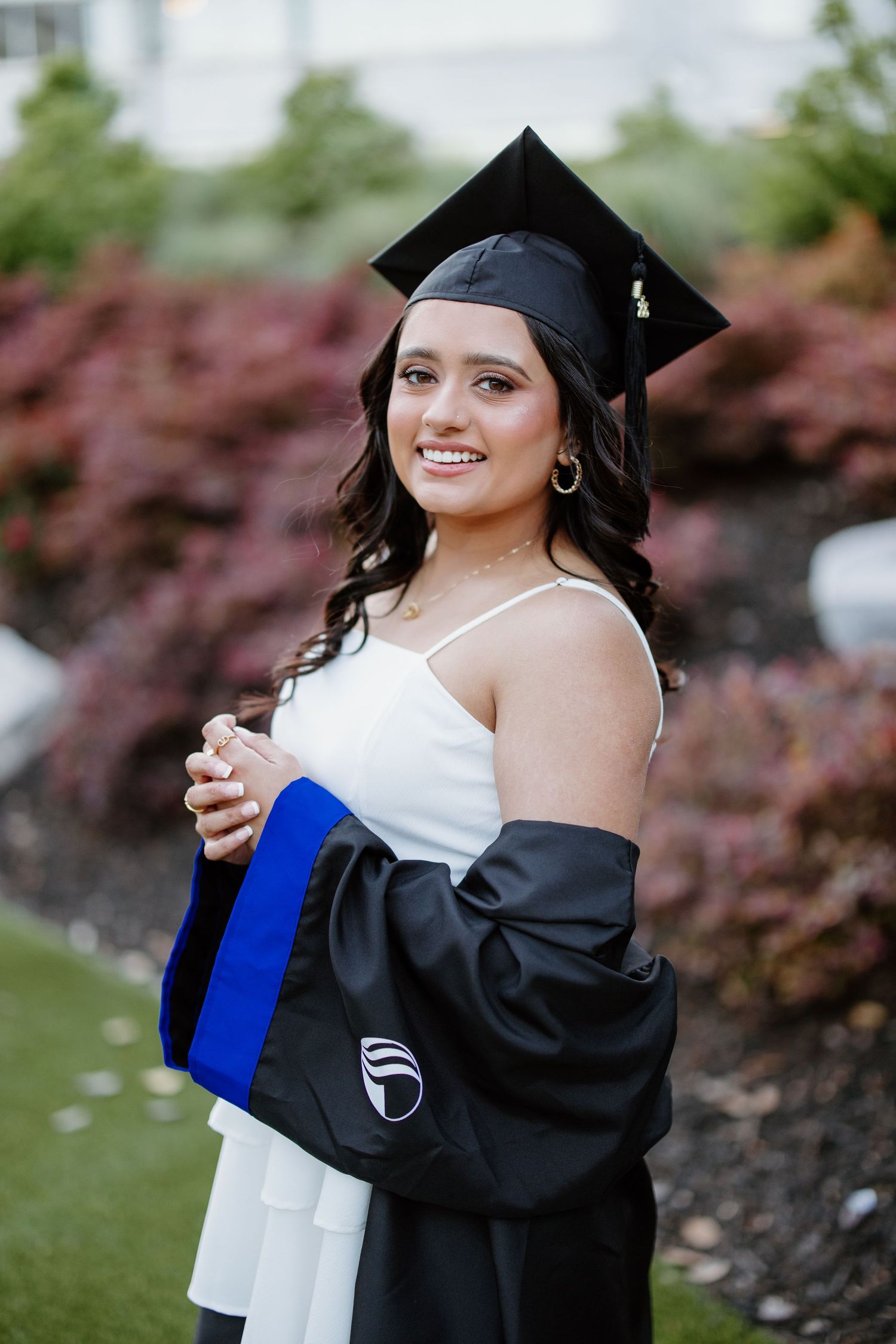 Graduate in cap and gown posing with diploma against landscaped background.