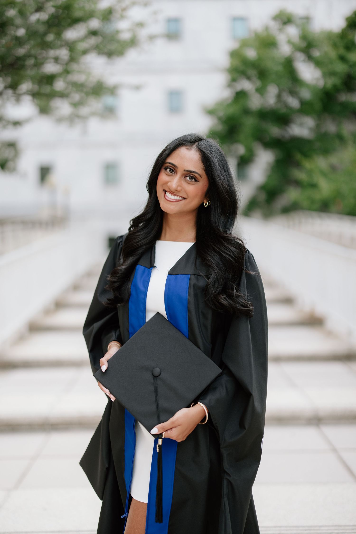 Smiling graduate in black gown and blue sash holds cap outdoors on campus steps.