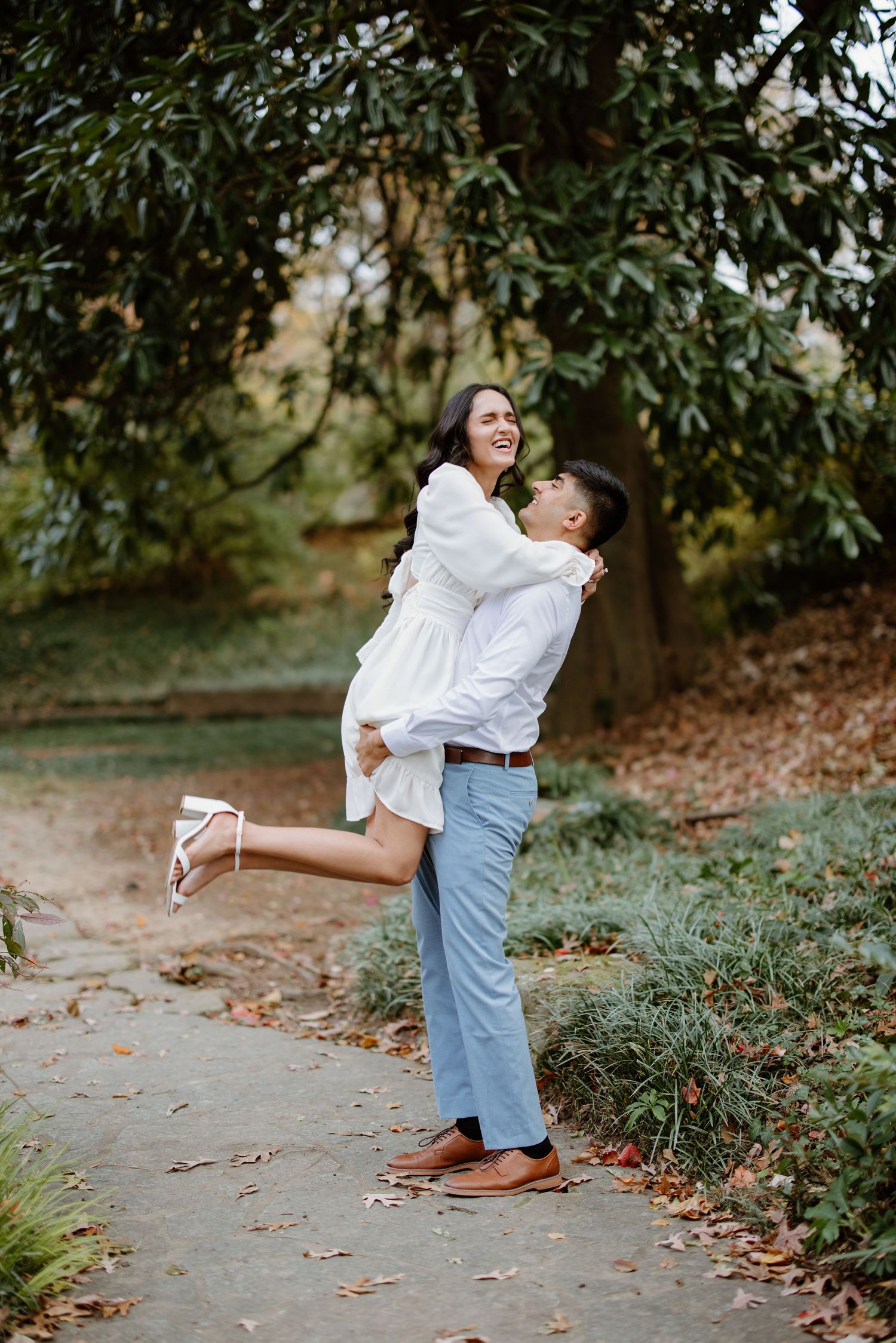 A romantic couple in casual attire shares an intimate embrace while standing on a leaf-covered pathway in a garden setting.
