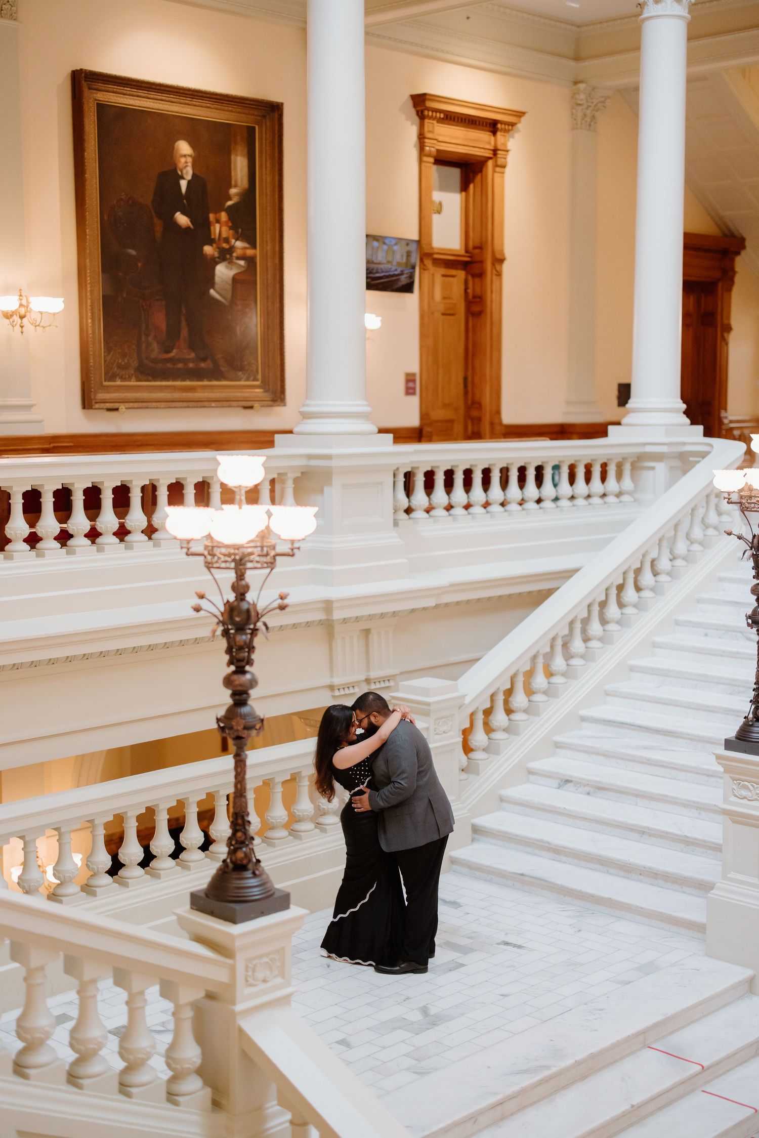 A couple embraces on an elegant marble staircase with white balustrades and ornate decor in a historic building.