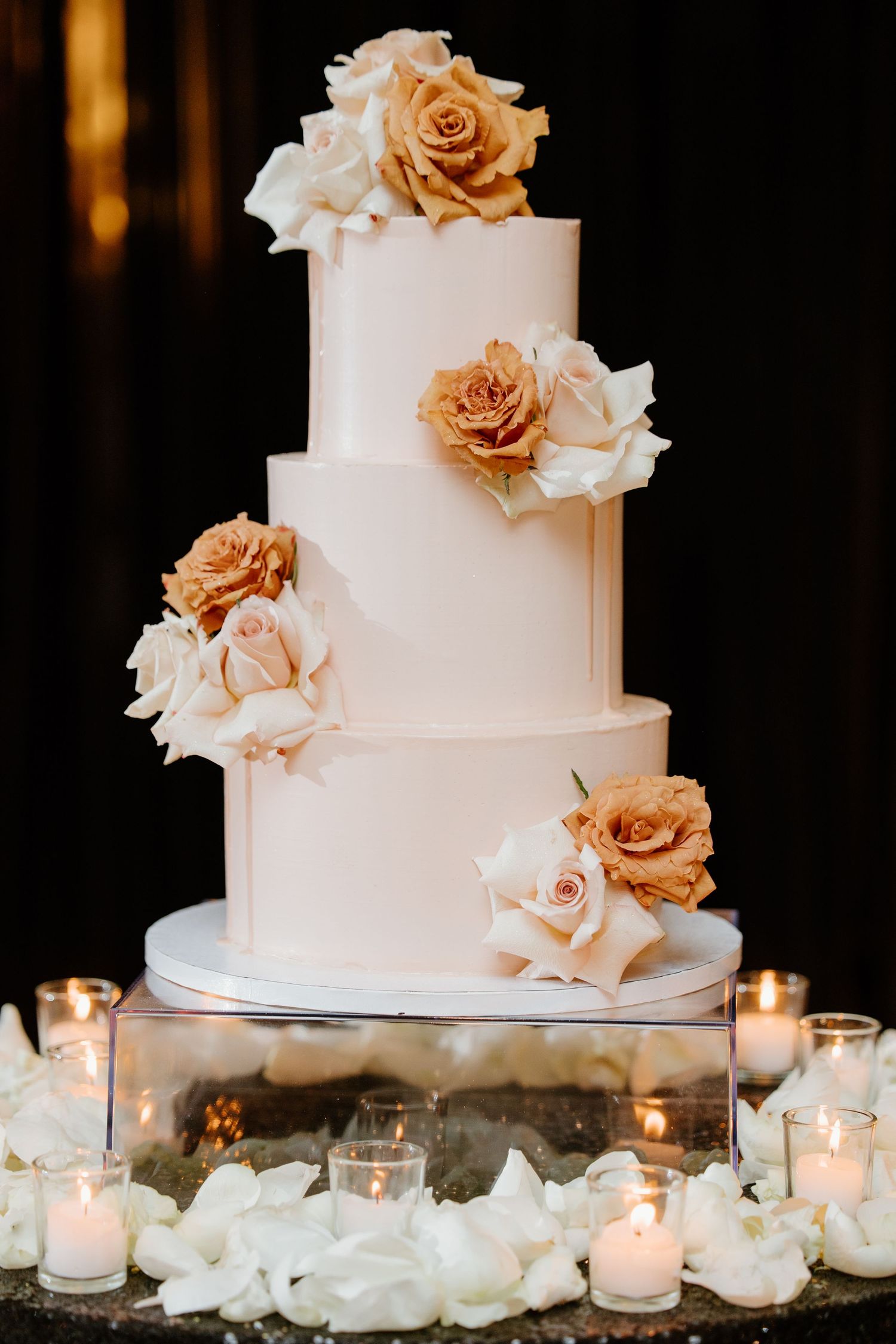 An elegant white wedding cake decorated with cascading champagne-colored roses and surrounded by glowing votive candles.