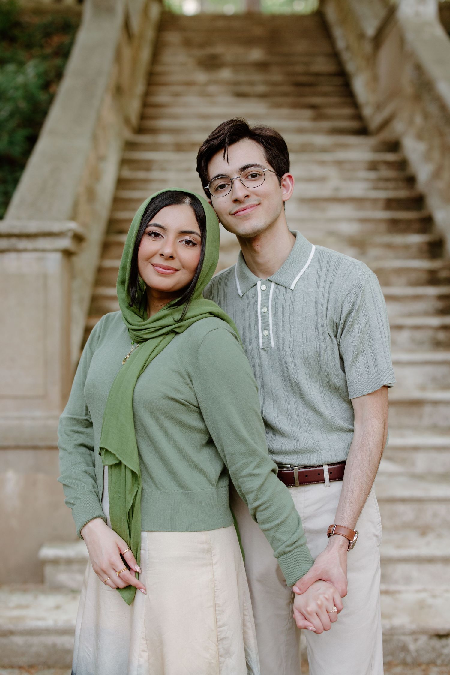 A couple stands together holding hands on stone steps, with one person wearing a green hijab and the other in glasses.