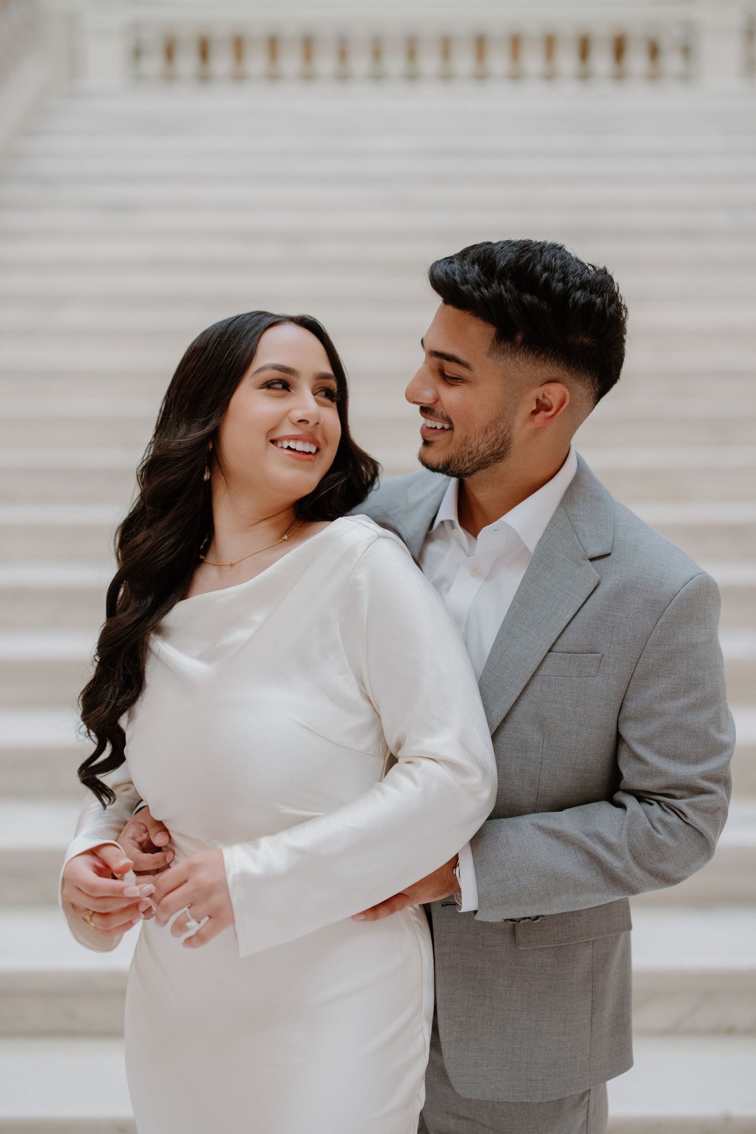 A couple shares a romantic moment while dressed in white and grey formal attire by stone steps.