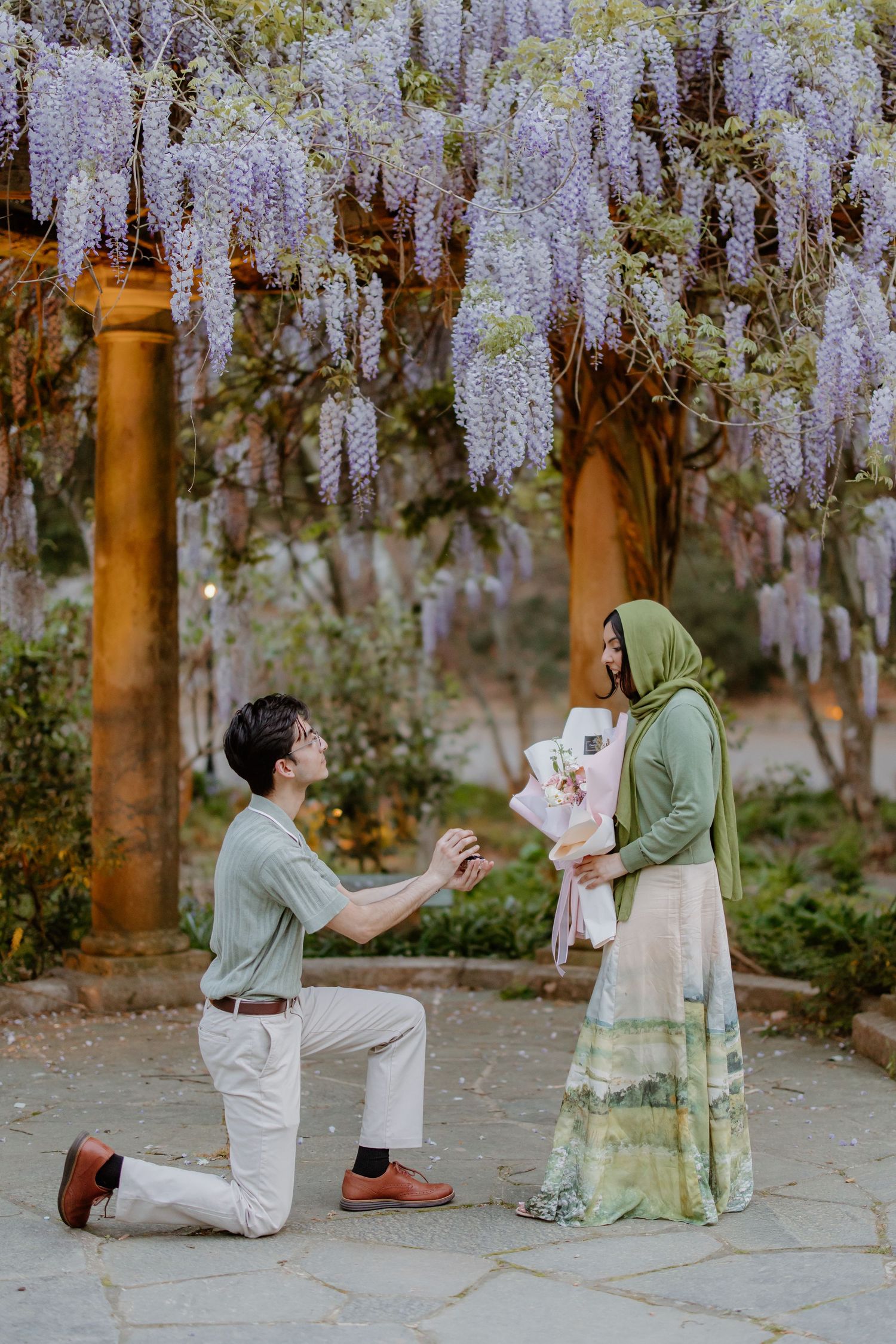 A couple share a romantic proposal moment under blooming wisteria trees in a garden.