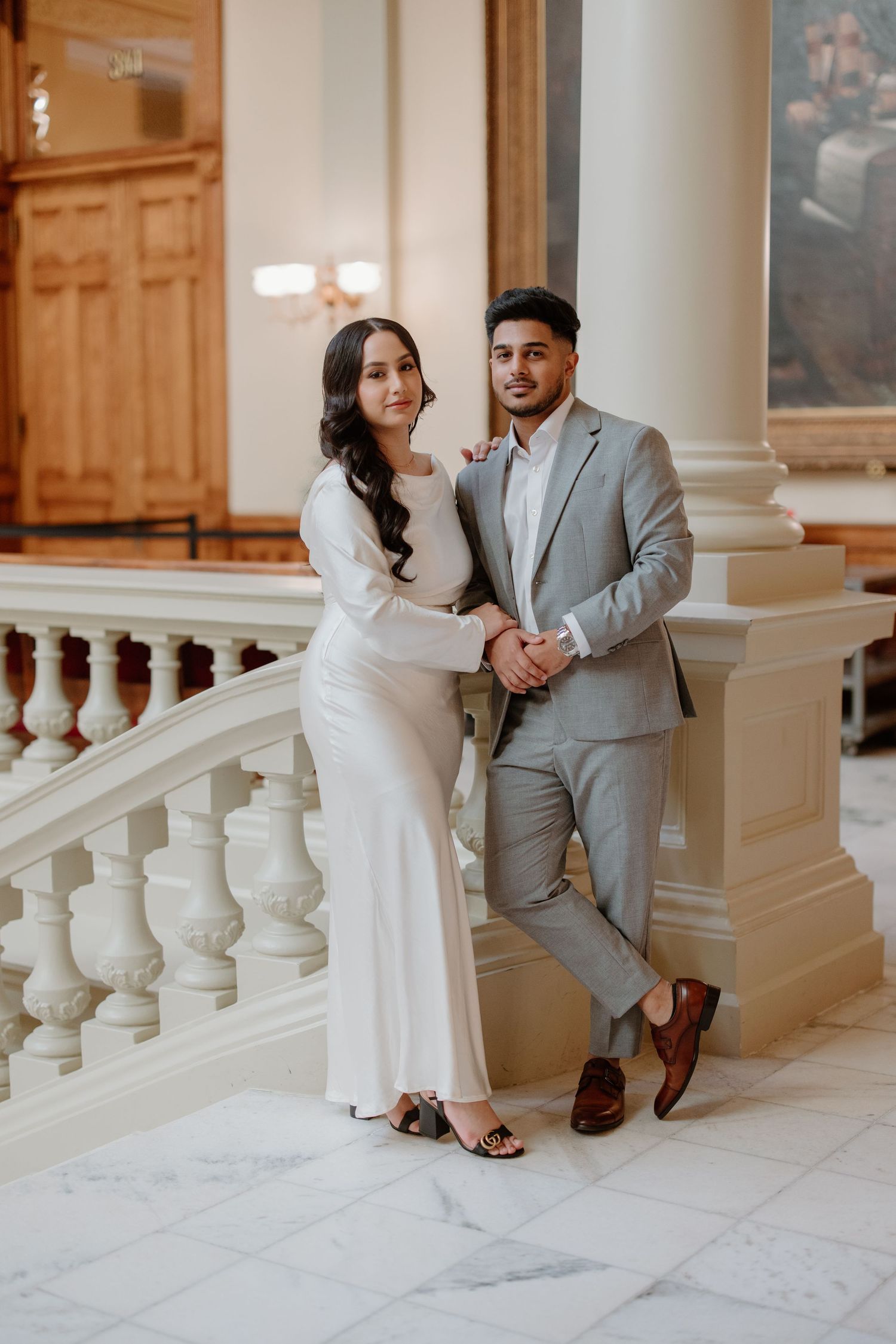 Romantic couple poses together on elegant white marble staircase with ornate balusters in formal wedding attire.