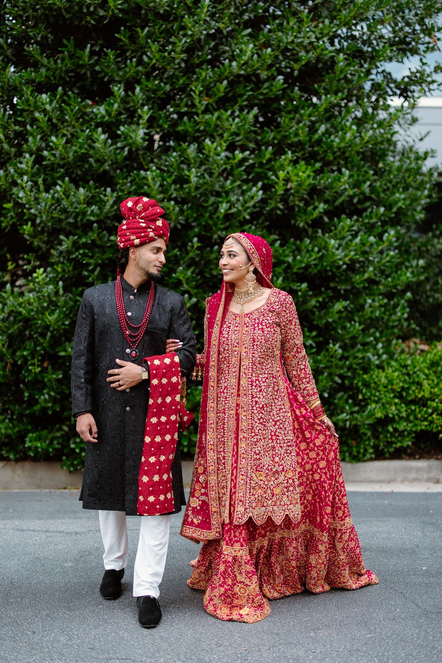 A couple in traditional South Asian wedding attire with red and black formal dress walking together outdoors.