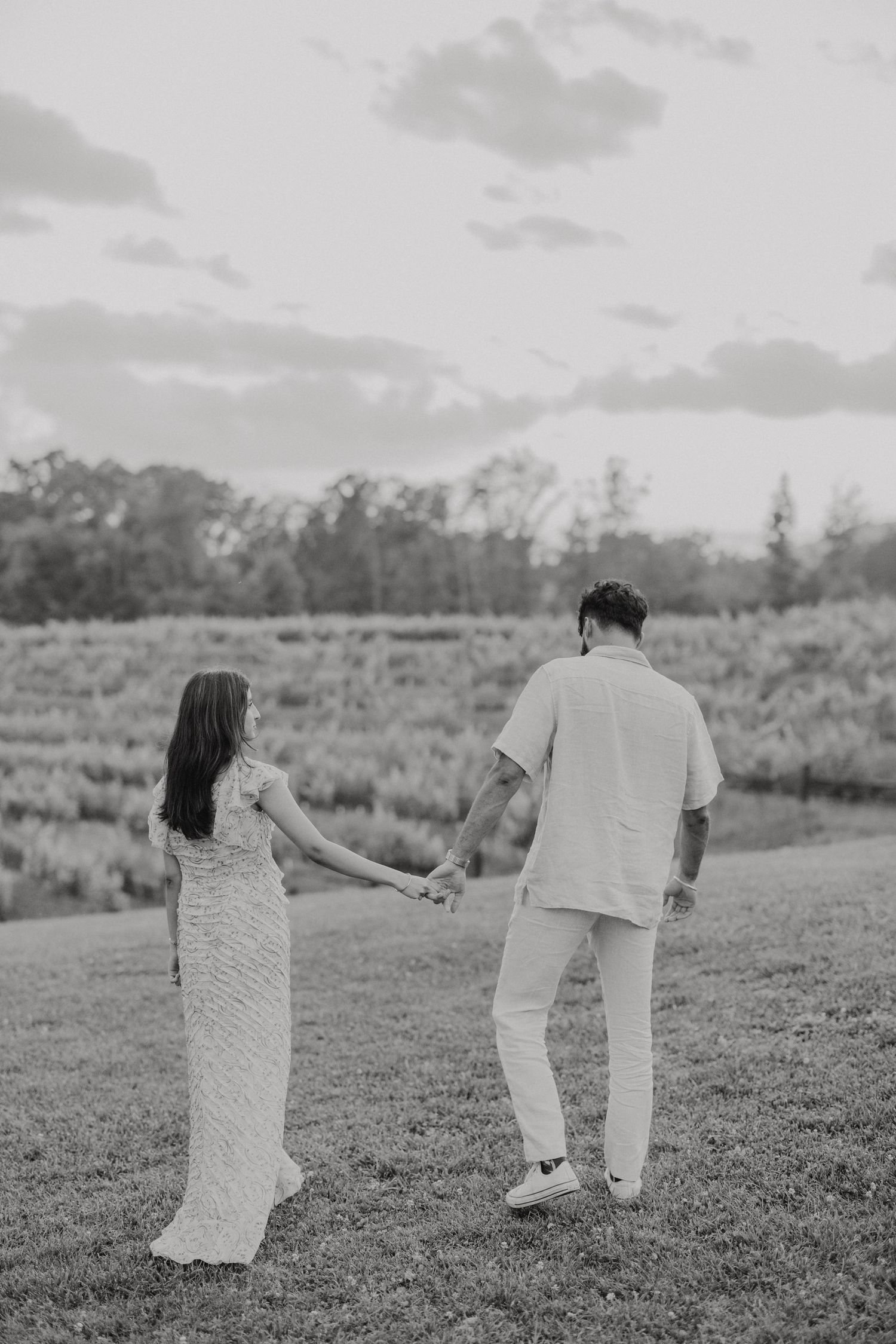 A couple holds hands in a grassy field at sunset while wearing matching white outfits.