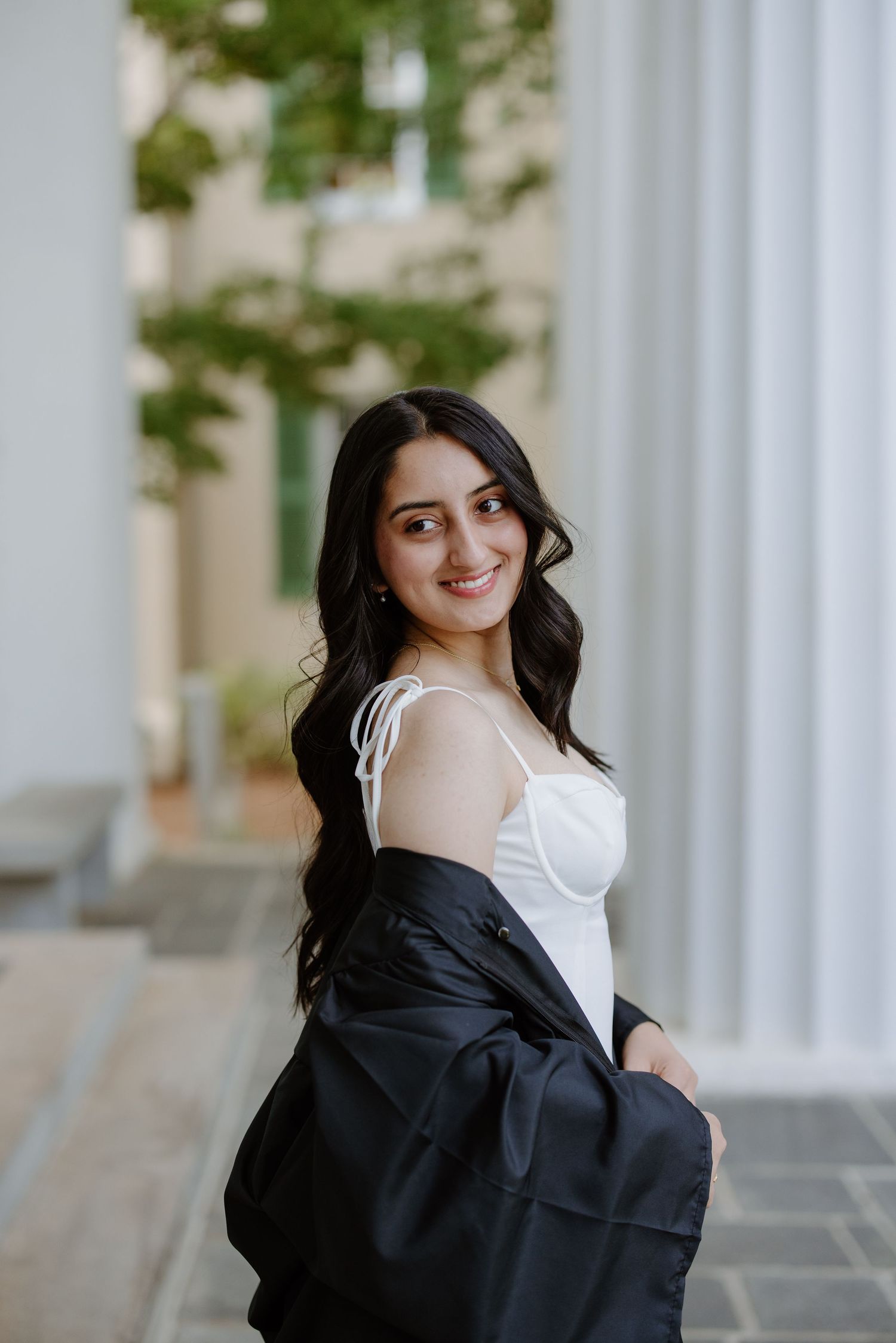 Young woman with long dark hair smiles over shoulder, wearing white top and black graduation gown outdoors.